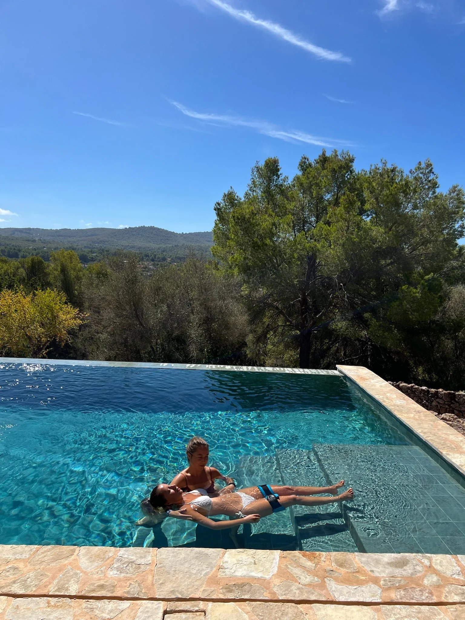 Two women relaxing in a swimming pool outdoors on a sunny day, with trees and hills in the background and a clear blue sky.