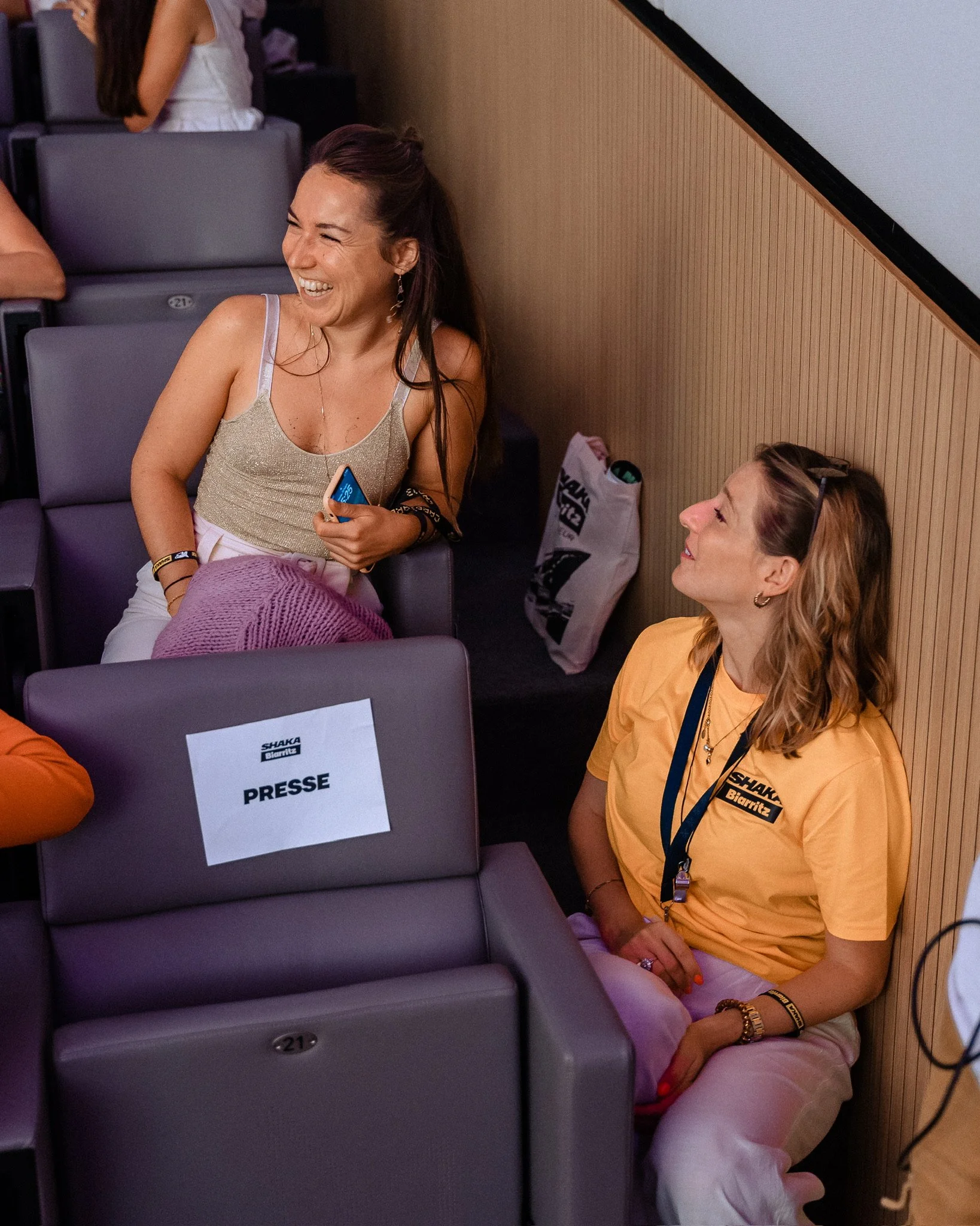 Two women sitting in chairs, sharing a laugh. One woman is sitting in an aisle seat, holding a phone and wearing a tank top and white pants. The other woman, sitting on the floor, wears a yellow t-shirt with a lanyard, and glasses on her head. There are signs on some of the chairs, with one reading 'Presse', indicating a media or press event.