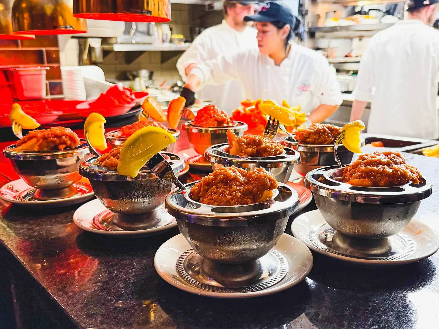 Multiple small bowls of fried chicken with lemon wedges on a dark counter in a restaurant kitchen, with chefs preparing food in the background.
