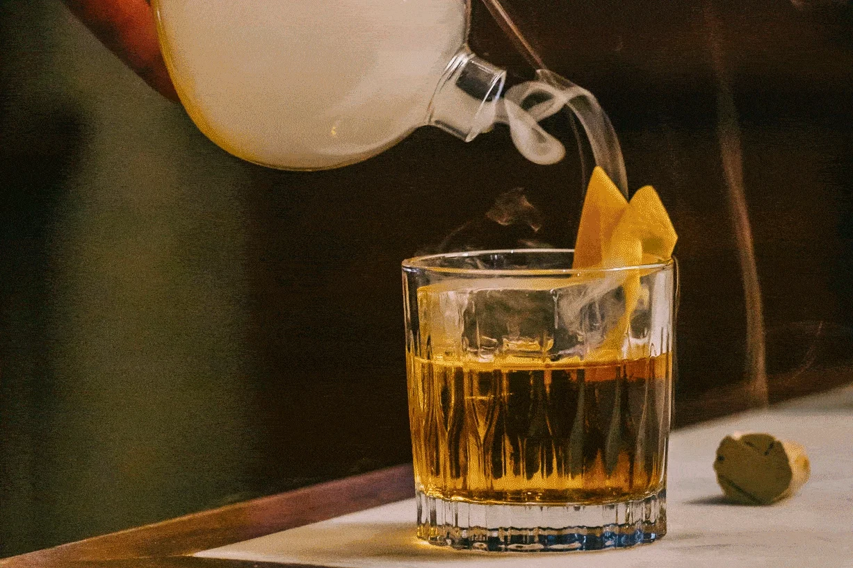 A cocktail with ice and lemon slice, with a teapot pouring smoke into the glass, on a wooden surface.