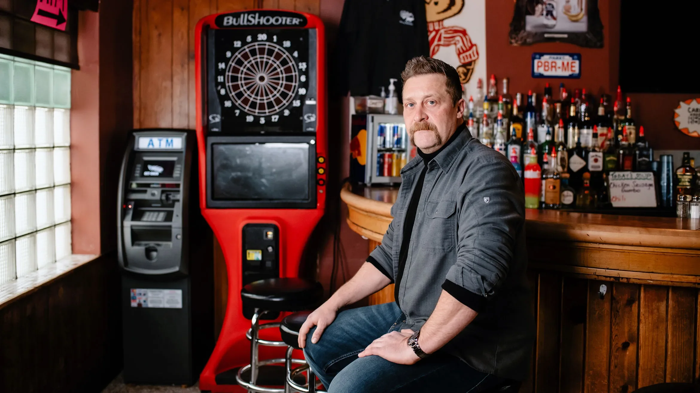 A man with a beard and mustache sitting at a bar, wearing a gray button-up shirt with rolled-up sleeves and blue jeans, with a dartboard, an ATM machine, and shelves with various bottles of alcohol in the background.