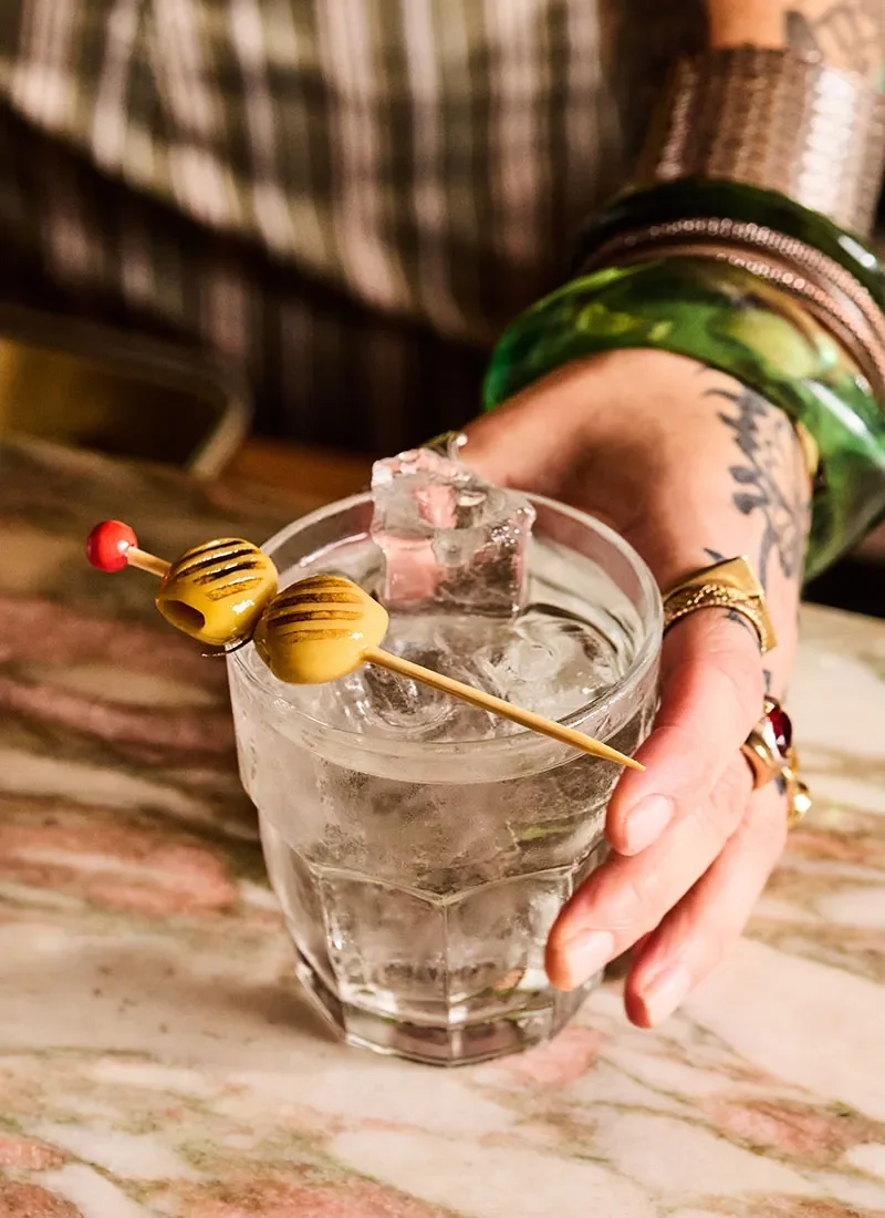 A hand with rings and bracelets holding a glass of clear liquid with ice and a large ice cube, garnished with a toothpick skewering yellow candies.