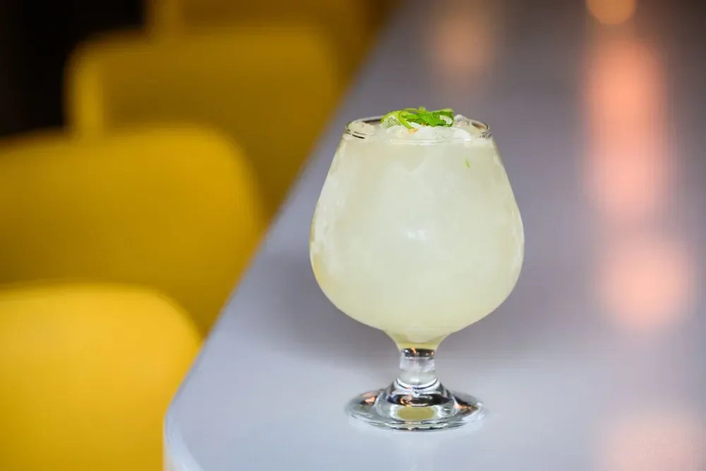 A pale yellow cocktail garnished with a slice of lime or lemon, served in a stemmed glass, on a bar or counter.