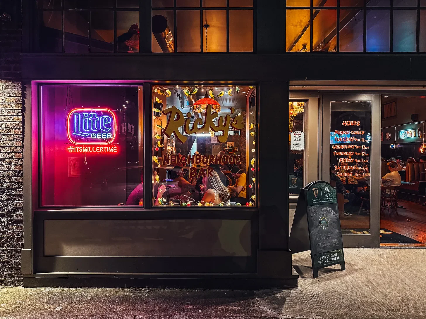 The storefront of Ricky's Neighborhood Bar at night, with neon signs for Miller Lite and other advertisements on the windows. Inside, people are sitting at tables, some wearing masks, and the interior is warmly lit with decorations. Outside, a chalkboard sign displays menu or event details.