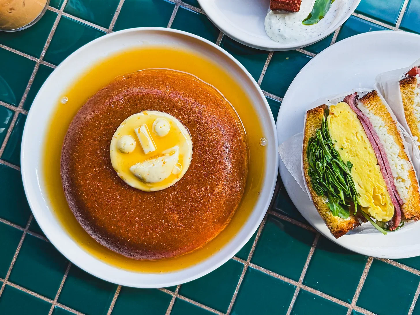 A large pancake with butter and syrup, served with a smiley face. Next to it is a sandwich with eggs, lettuce, ham, and cheese.