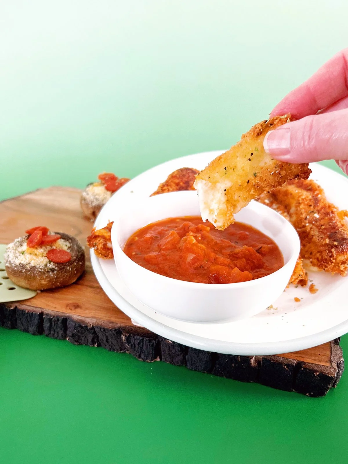 Hand dipping a crispy deep-fried mozzarella stick into marinara sauce beside breaded mozzarella slices and stuffed mushrooms on a rustic wood serving board against a retro green background.