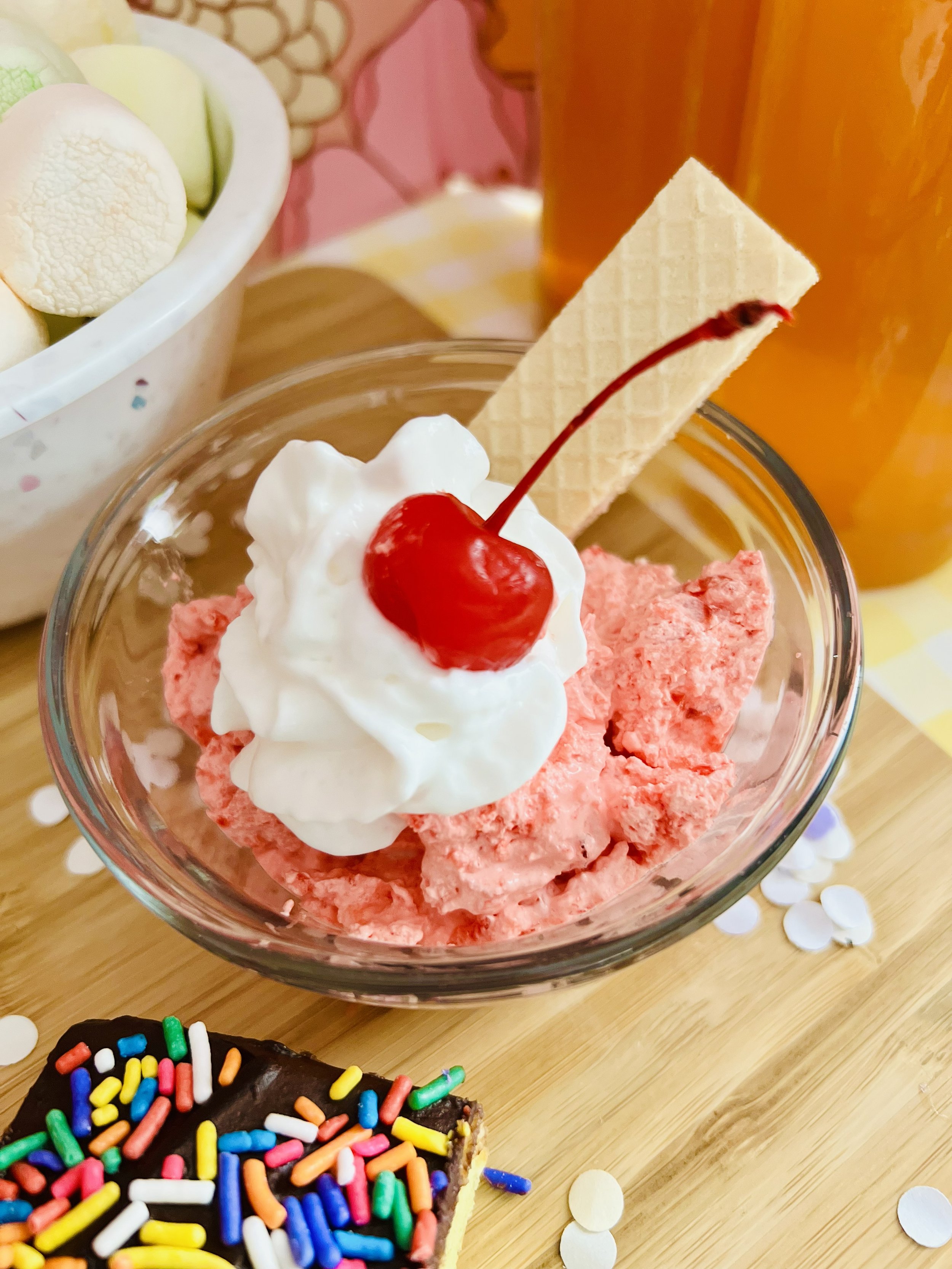 Maraschino Cherry Cheesecake Fluff served in a vintage amber dessert bowl, topped with crushed graham crackers and a maraschino cherry. A bowl of pastel marshmallows 