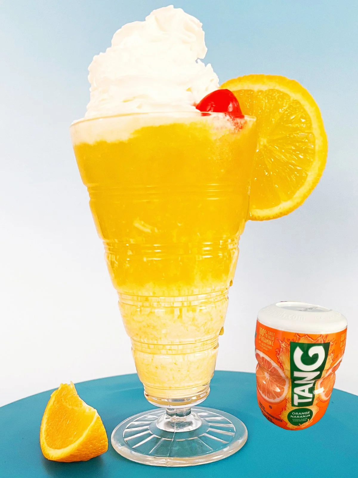tang Orange Tang fizzy soda in a vintage-style glass topped with whipped cream, a cherry, and an orange slice on a blue background