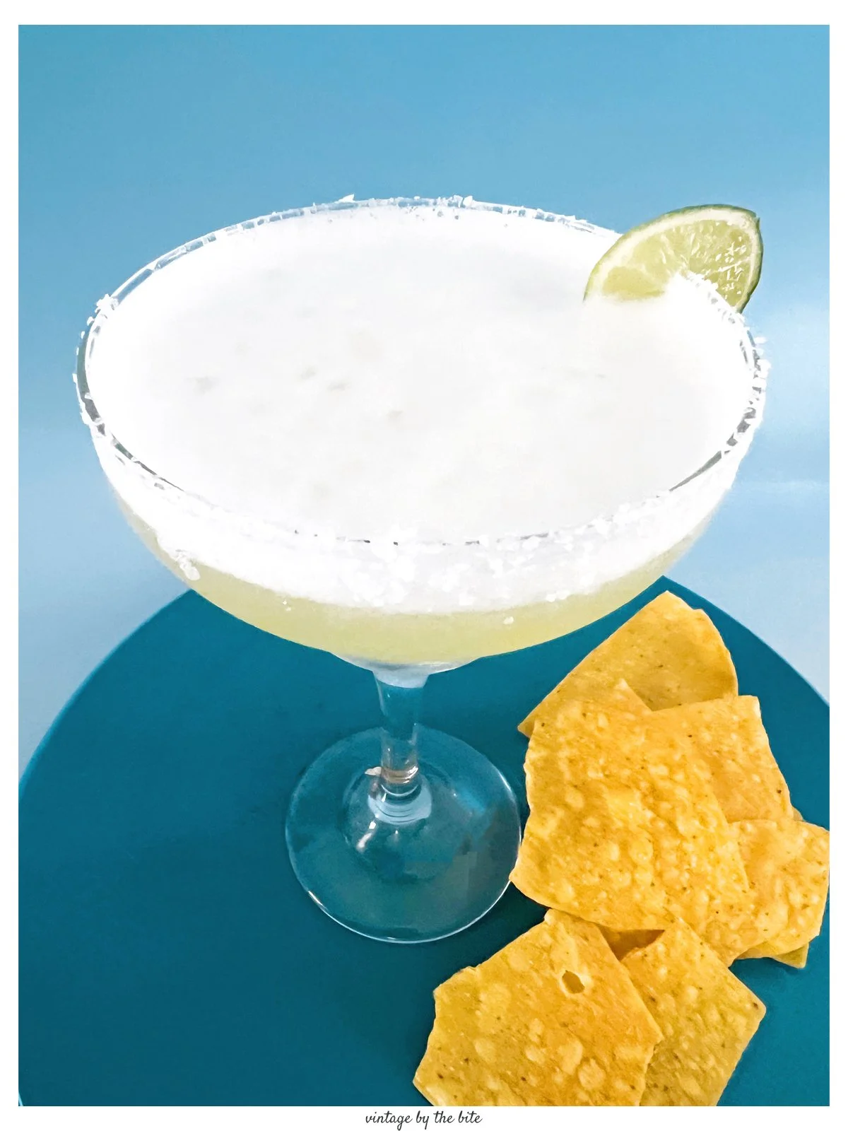 Close-up of a frothy 1970s-style margarita with a salted rim and lime wedge, served in a wide glass on a teal plate with tortilla chips.