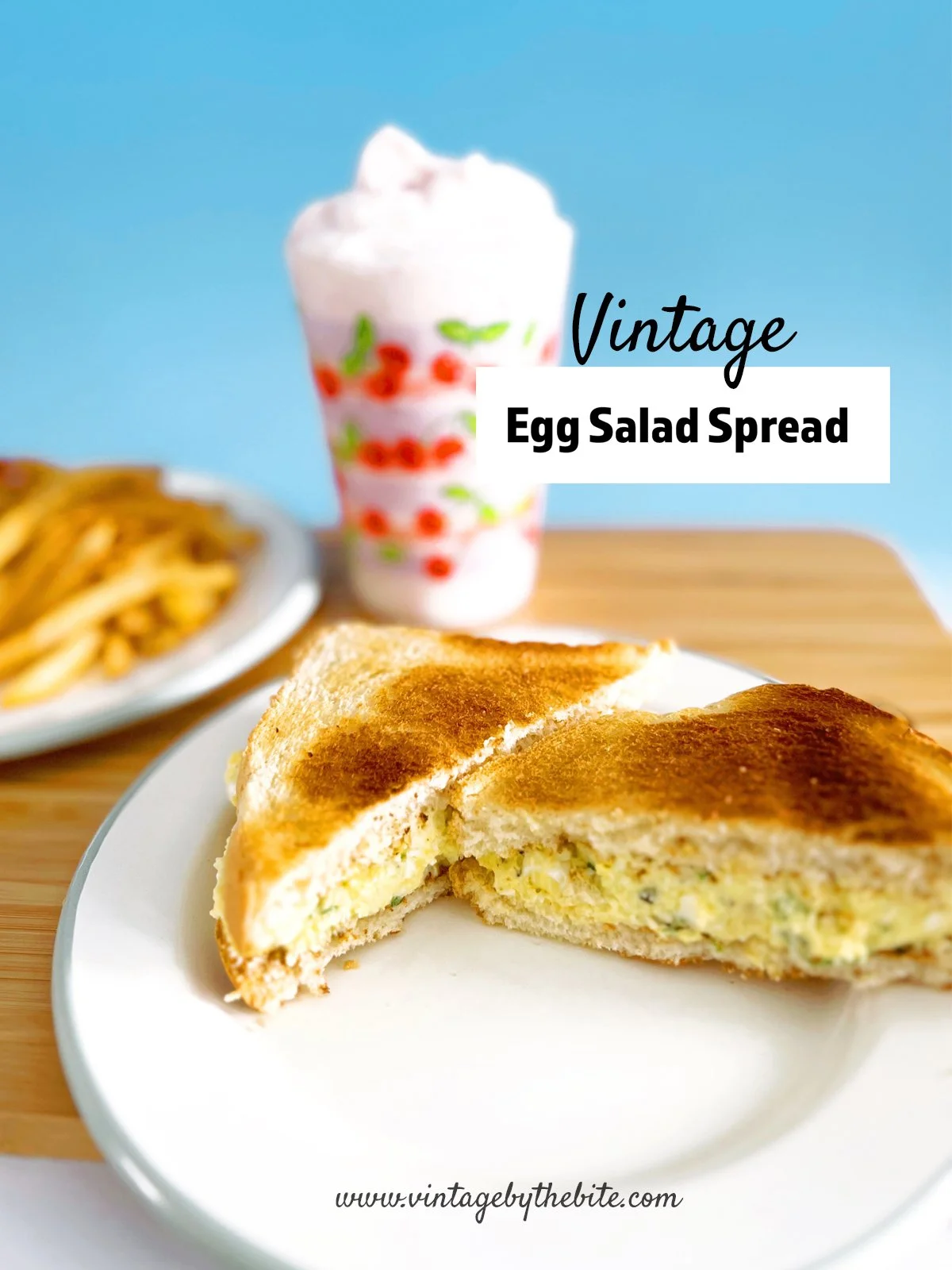 A toasted vintage-style egg salad sandwich cut into triangles sits on a white plate on a wooden table. The creamy egg salad filling is visible between slices of golden toasted bread. In the background, a plate of French fries and shake