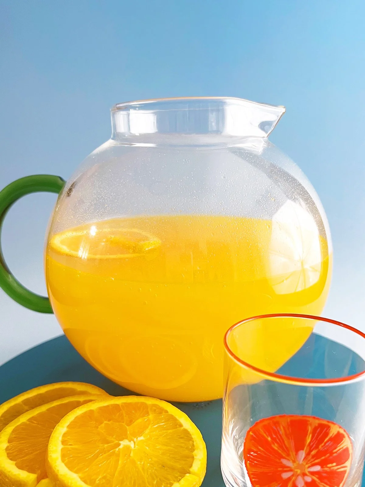 Glass pitcher of orange Tang soda with orange slices and a matching glass on a blue surface