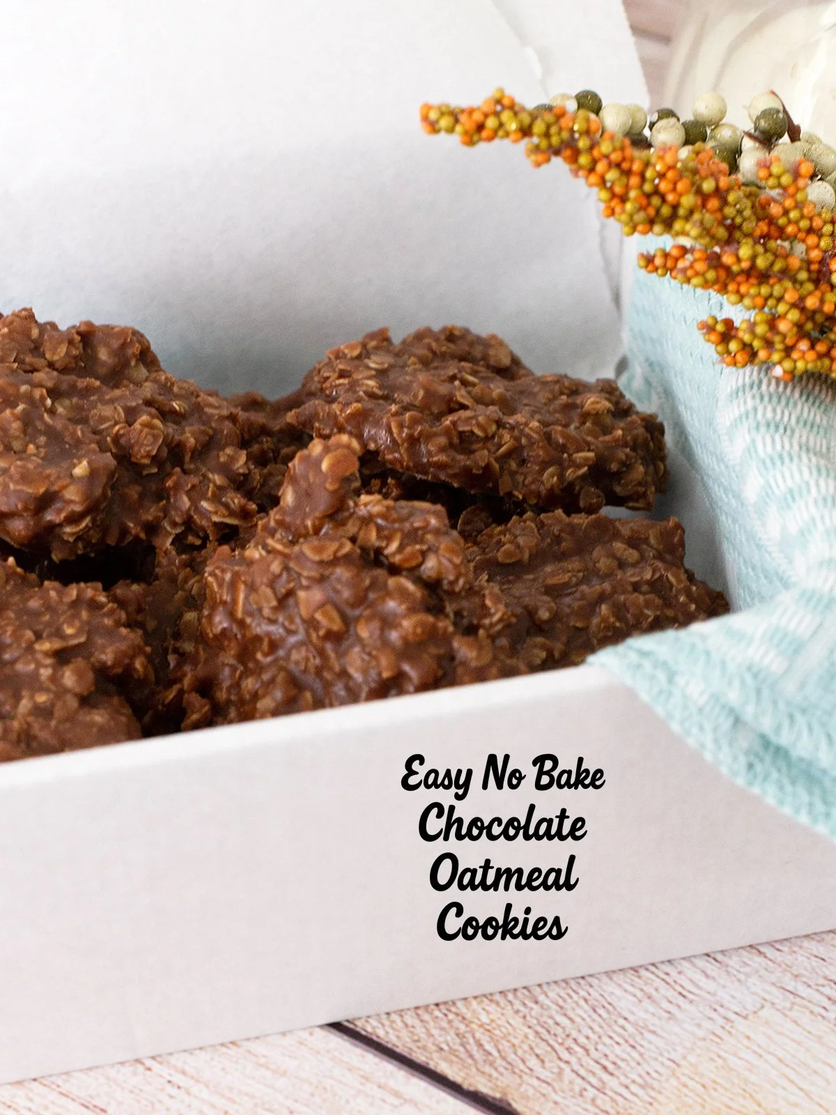 Close-up of a white box filled with easy no bake chocolate oatmeal cookies, topped with oats and cocoa for a chewy texture. A cozy fall display with a teal cloth and autumn-colored floral accent completes the rustic seasonal look.