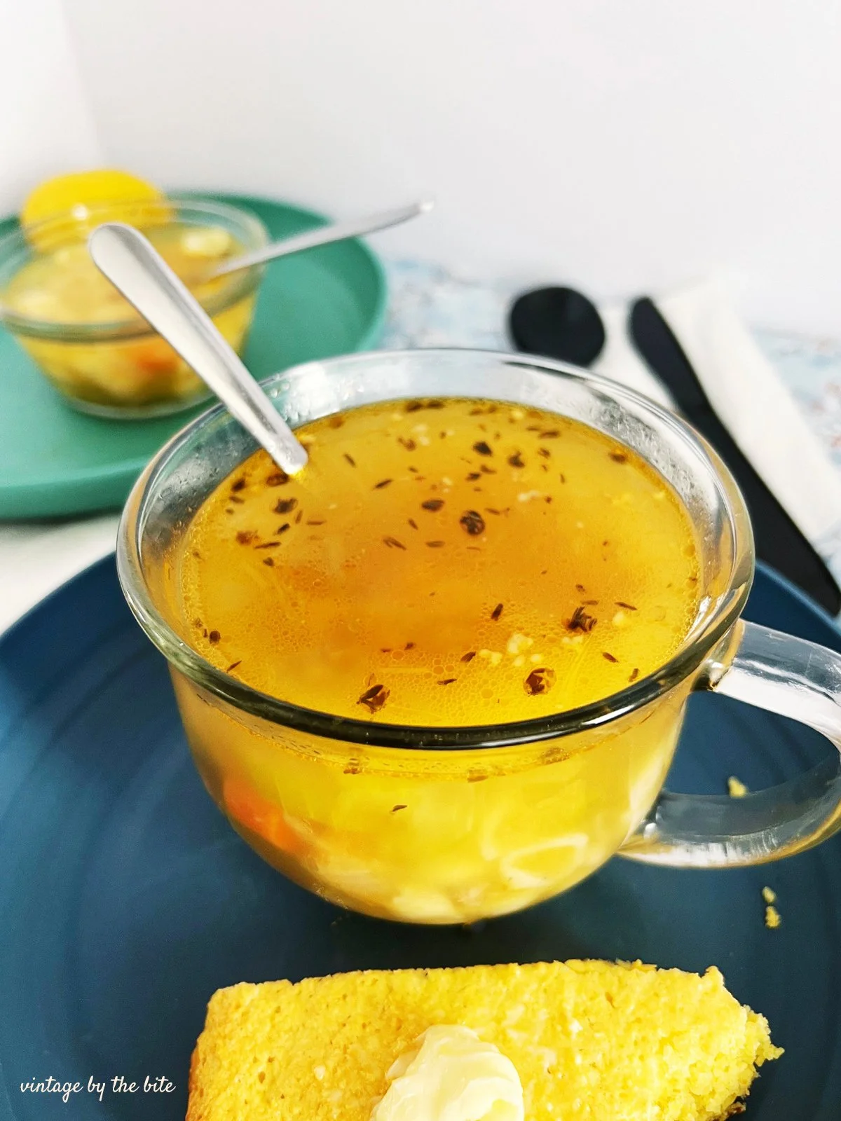 hero Mediterranean pasta soup served in a clear glass mug with lemon-infused broth, herbs, and shell pasta, paired with buttered cornbread on a blue plate in a retro 1950s kitchen setting.