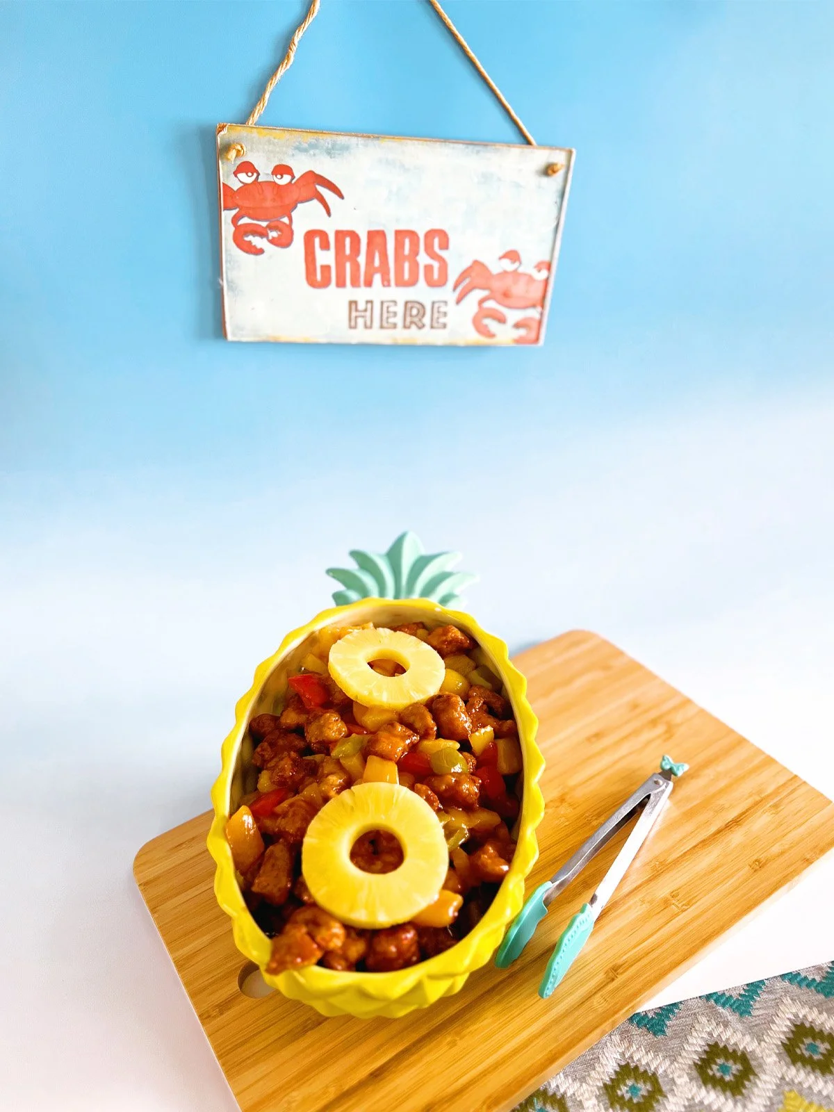 Sweet and sour chicken served in a yellow pineapple-shaped dish, topped with pineapple rings and mixed peppers, placed on a wooden board with serving tongs and a small vintage “Crabs Here” sign hanging in the background.