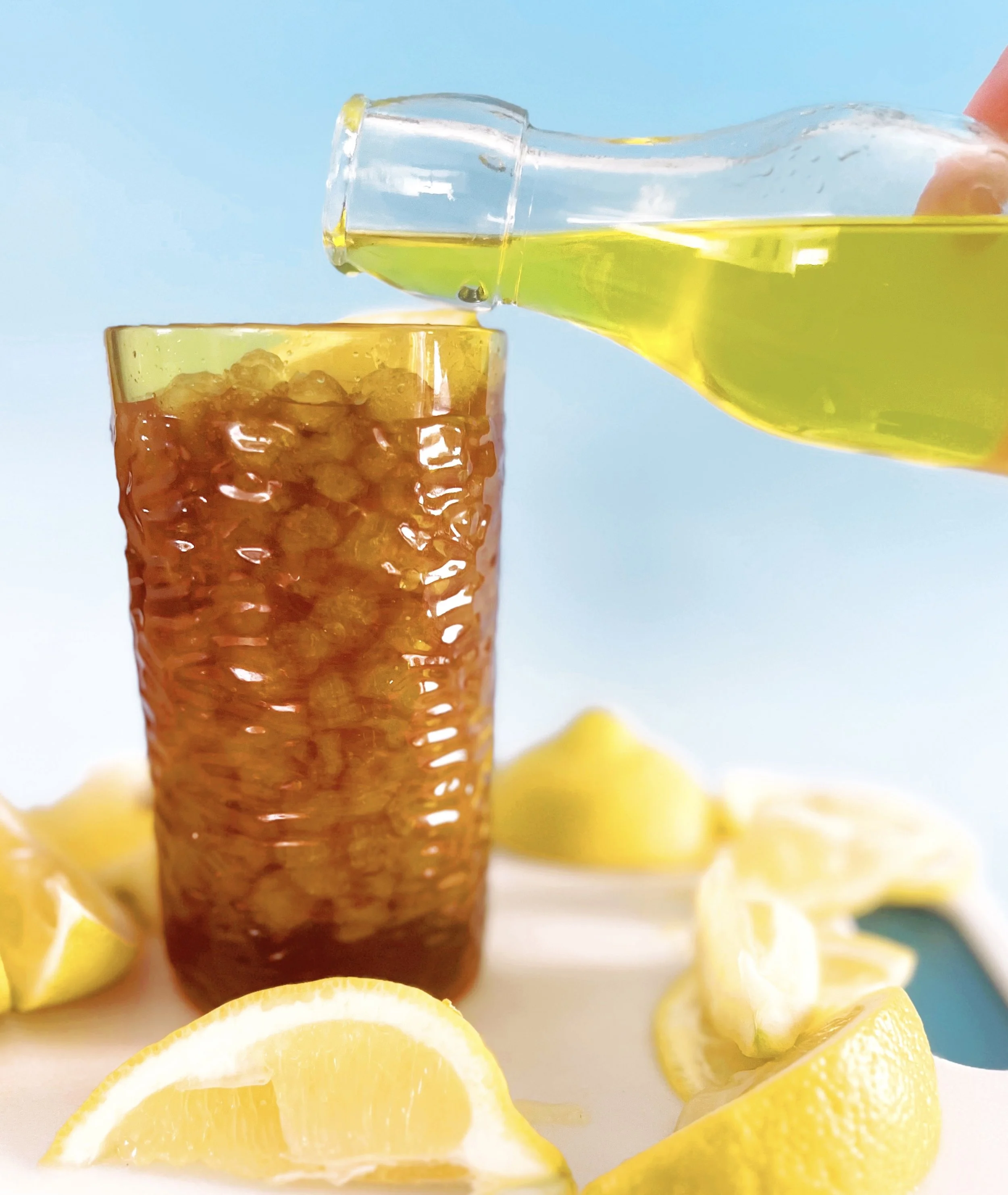 Bright yellow lemon syrup being poured from a small glass bottle into a tall glass of iced tea, surrounded by fresh lemon wedges on a light surface.