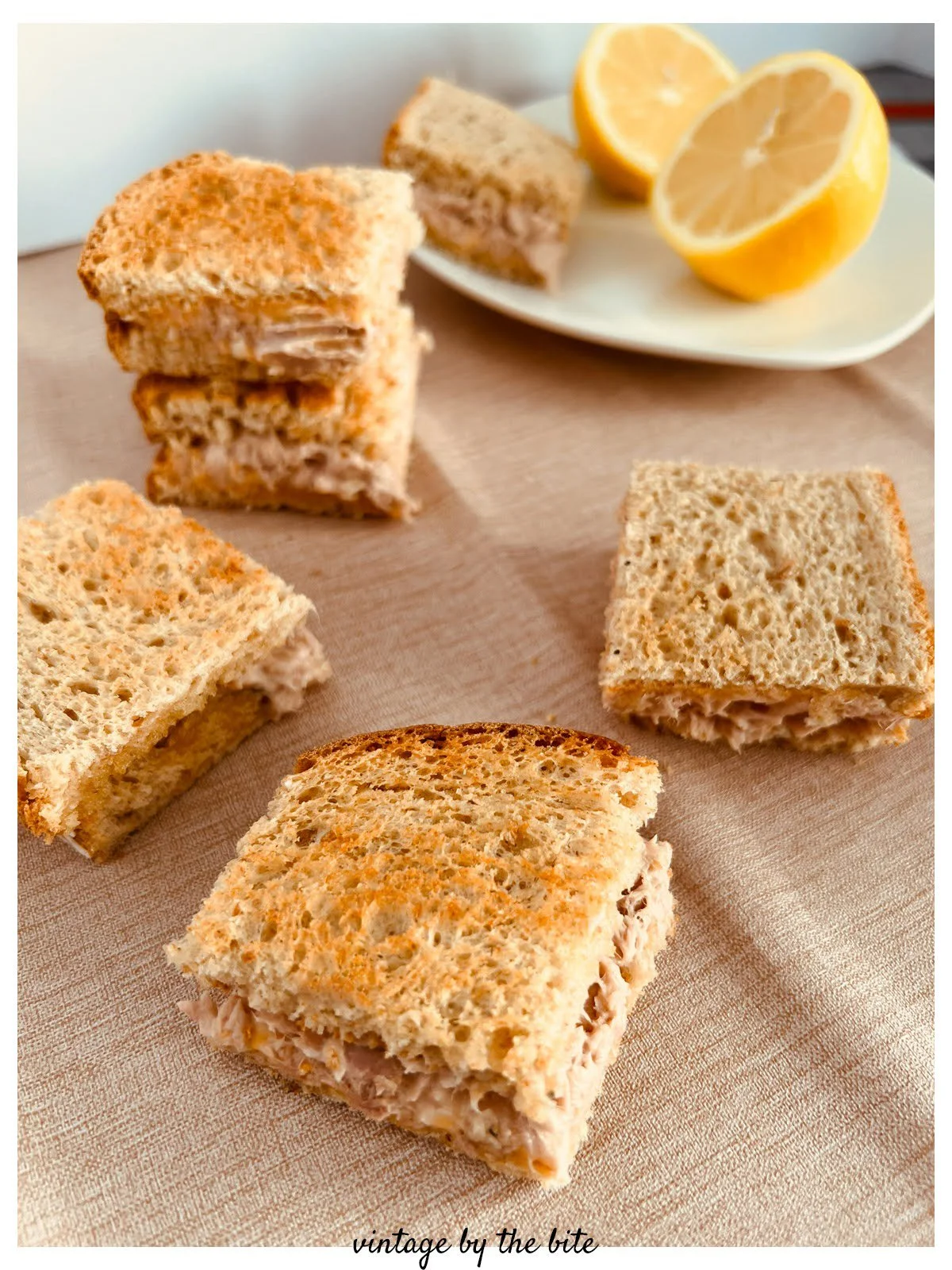 Cheesy tuna toasties cut into small squares and arranged on a tabletop with lemon halves in the background, featuring toasted white bread, creamy tuna filling, and melted Cheddar cheese, inspired by a 1958 Good Housekeeping recipe.