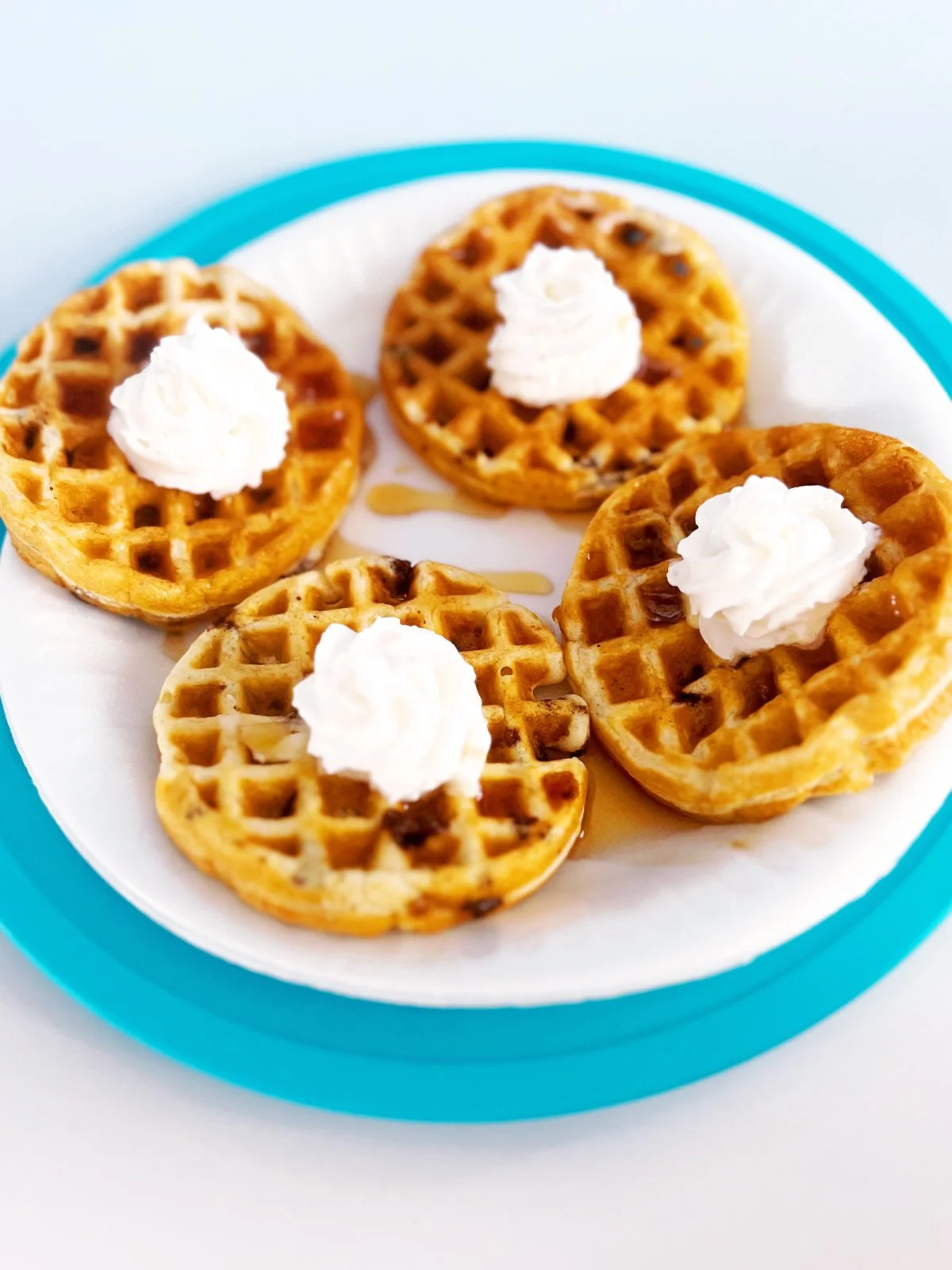 Four mini cappuccino chip waffles topped with whipped cream and chocolate chips, arranged on a white plate with a teal edge and lightly drizzled with syrup.