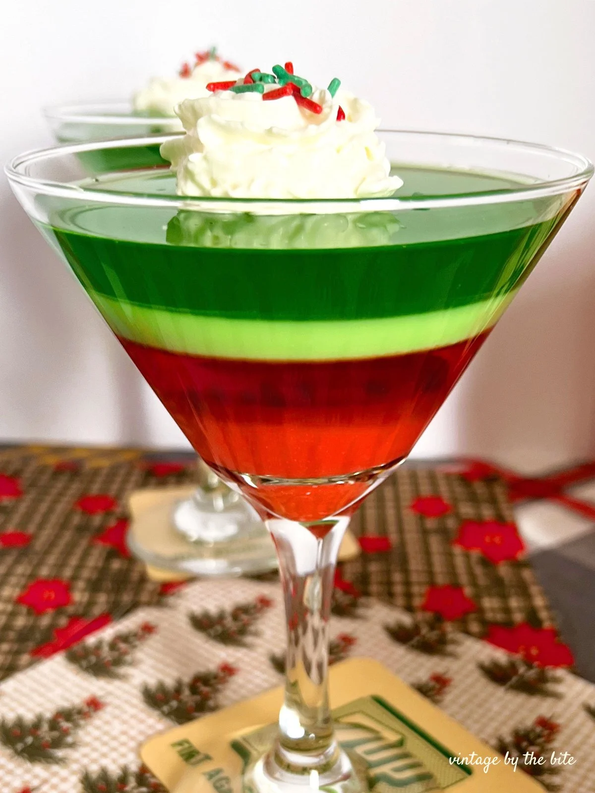 Close up Layered Holiday Ribbon Jell-O dessert served in martini glasses with red and green gelatin layers, topped with whipped cream and festive sprinkles on a vintage holiday table setting.