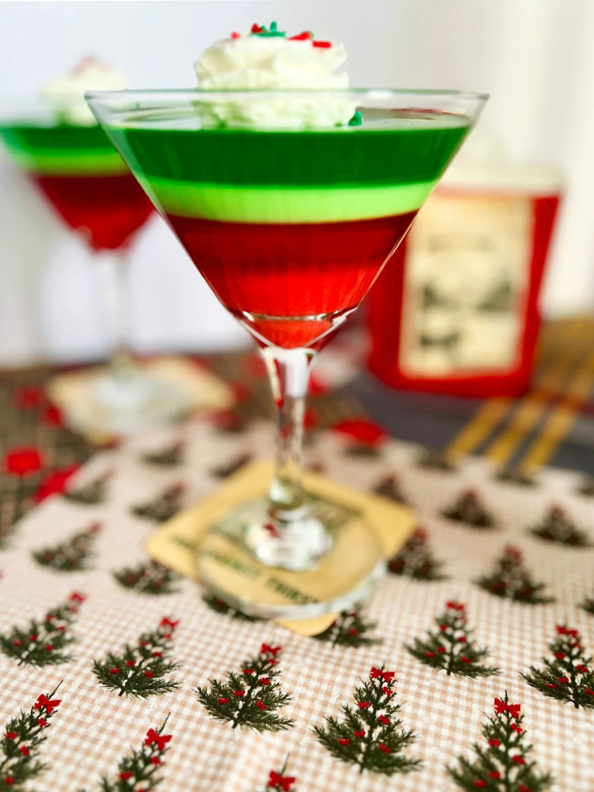 Holiday Ribbon Jell-O by the glass with layered red and green gelatin, served in a martini glass and topped with whipped cream and festive sprinkles on a vintage holiday tablecloth.