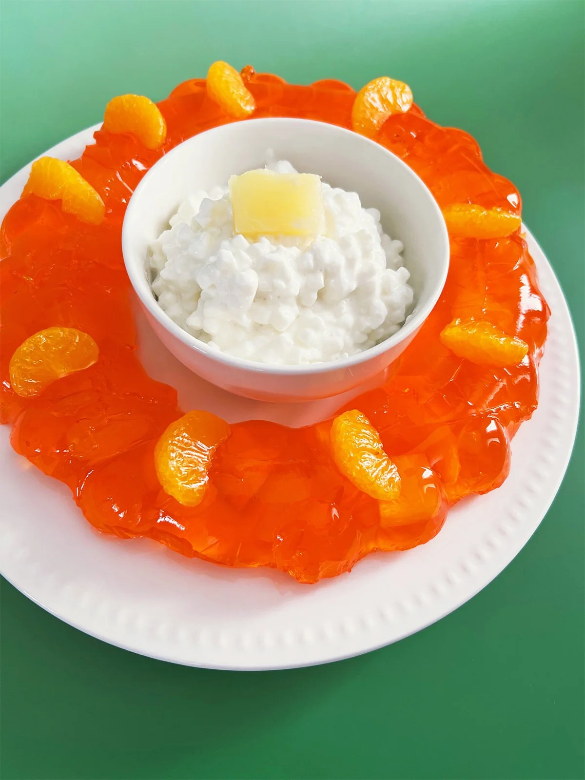 Bright orange mandarin pineapple Jell-O salad molded into a ring on a white plate, topped with mandarin slices and filled in the center with a bowl of cottage cheese and pineapple, styled in a 1960s retro entertaining theme