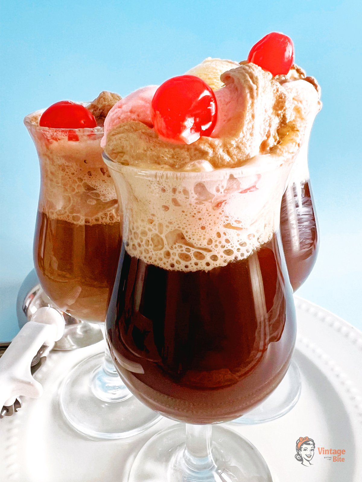 Three retro cola ice cream floats in soda fountain glasses topped with vanilla and chocolate ice cream and maraschino cherries.