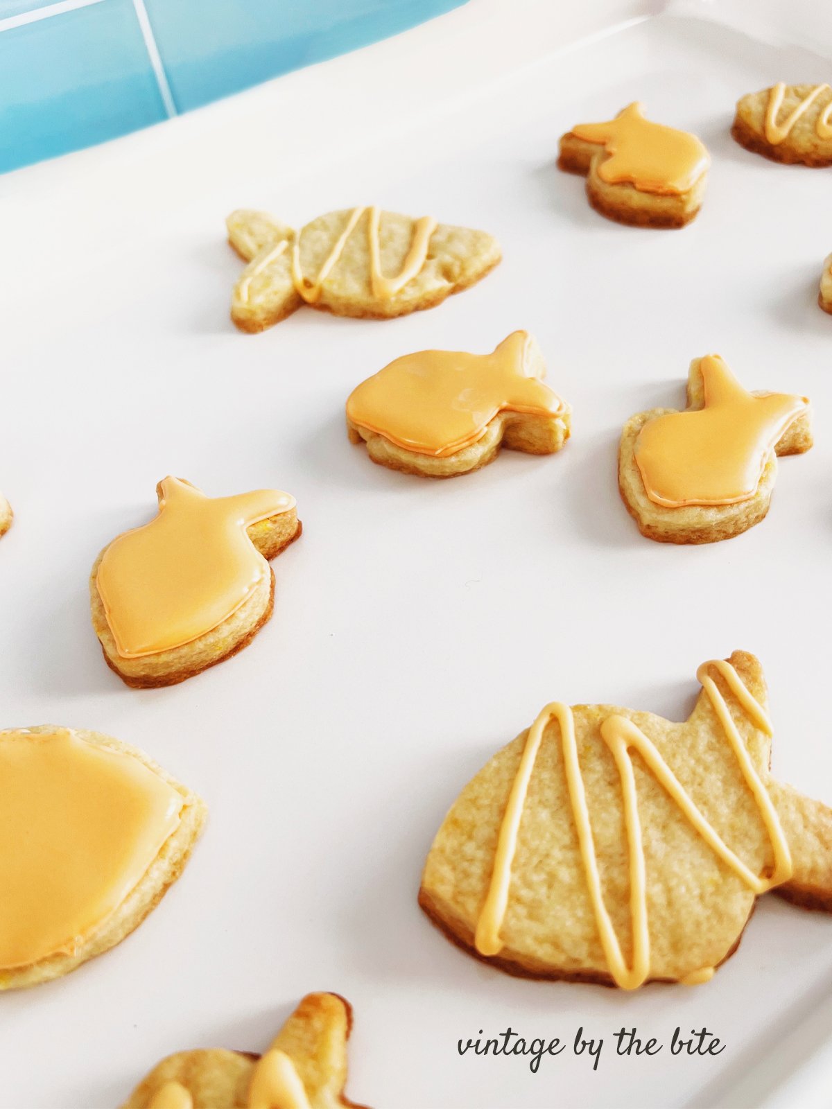 Goldfish-shaped sugar cookies decorated with a smooth orange icing and light drizzle, arranged on a white surface with a pale blue tiled background.