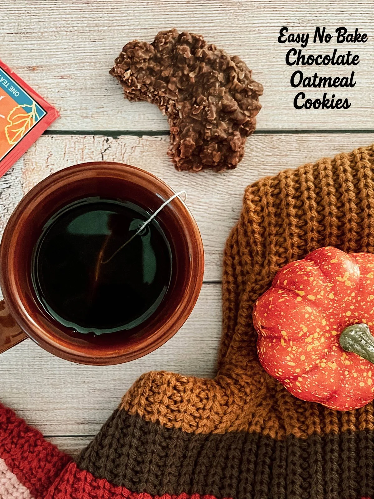 Overhead view of a cozy fall scene with a cup of hot tea, a half-eaten no bake chocolate oatmeal cookie, a knitted autumn sweater, and a small decorative pumpkin on a rustic wooden table