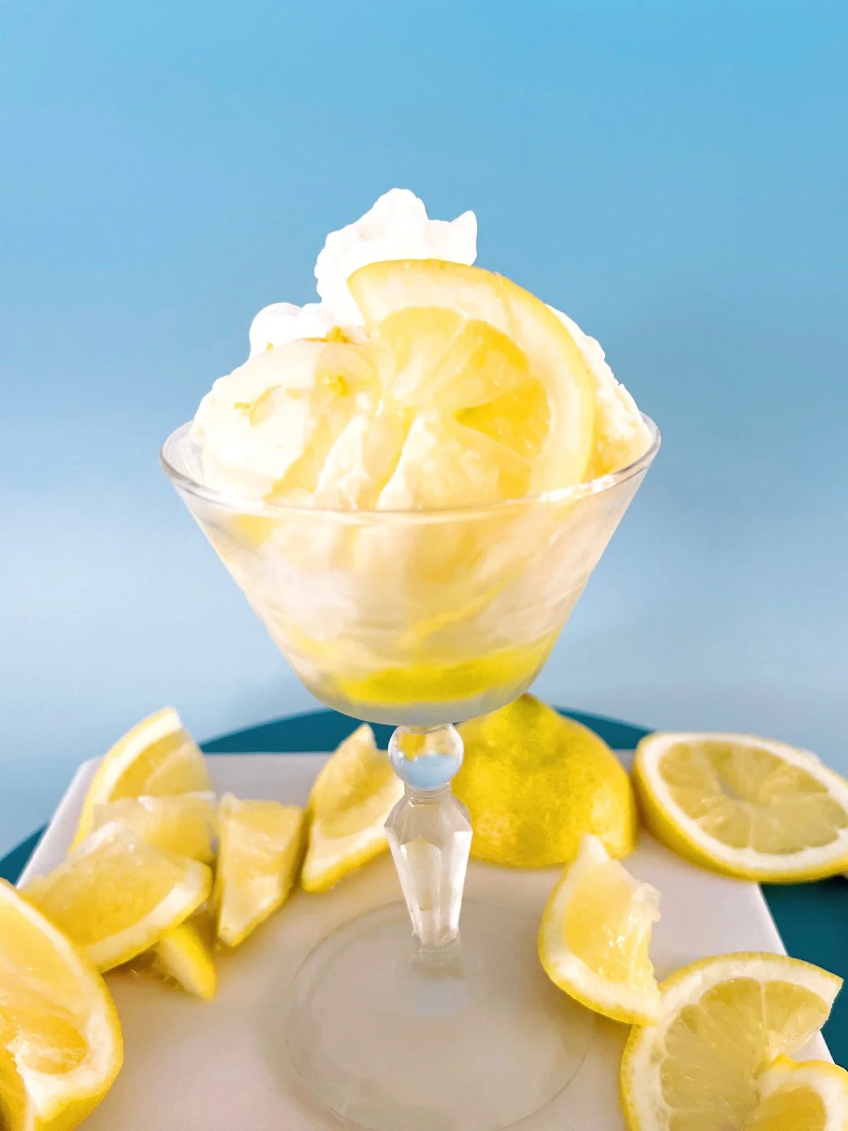 Small glass bottle filled with vibrant yellow lemon syrup, styled on a tray with fresh lemon slices against a soft blue background.