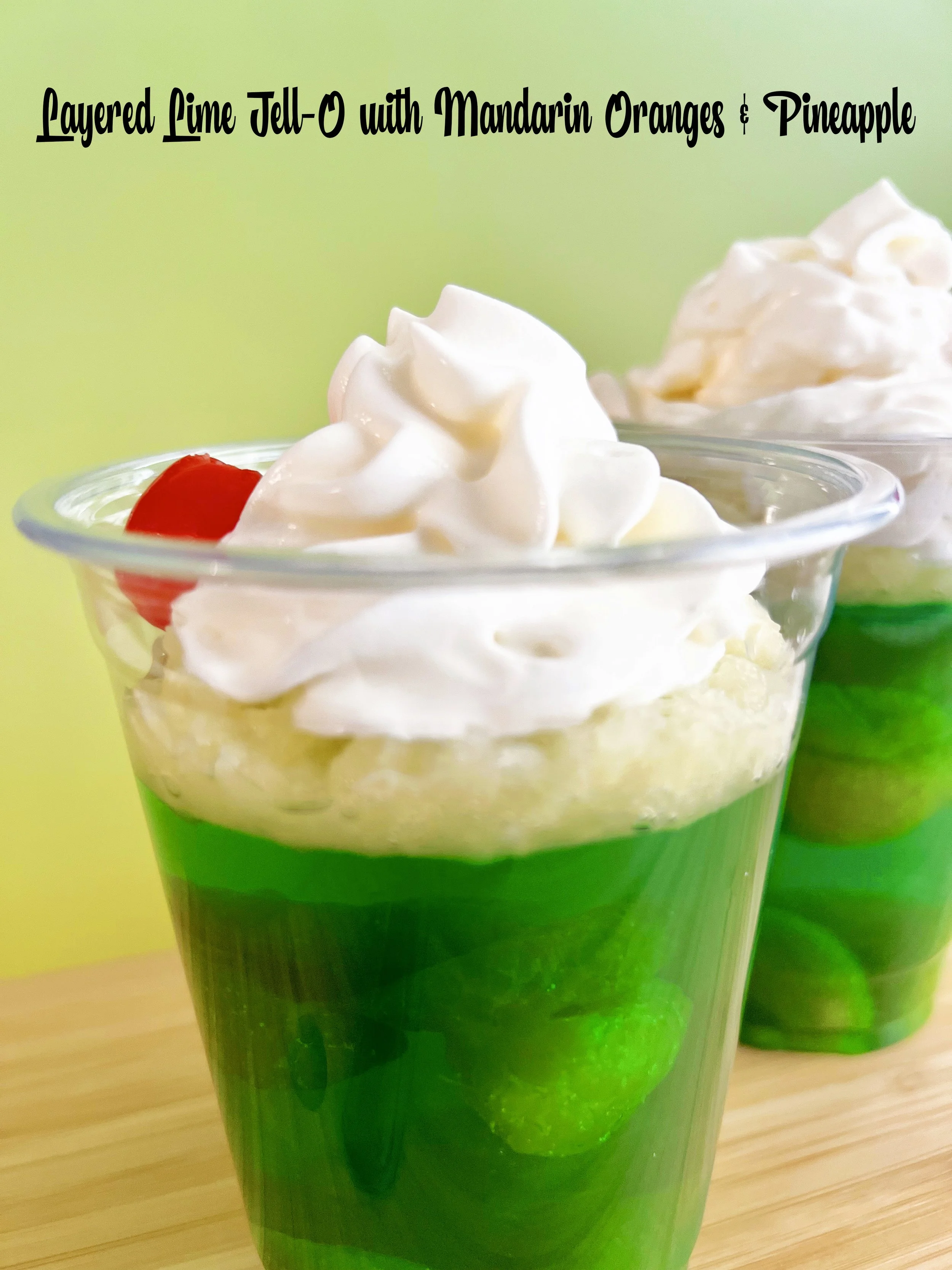 Layered lime Jell-O dessert in a clear cup with mandarin oranges, crushed pineapple, whipped cream, and a maraschino cherry