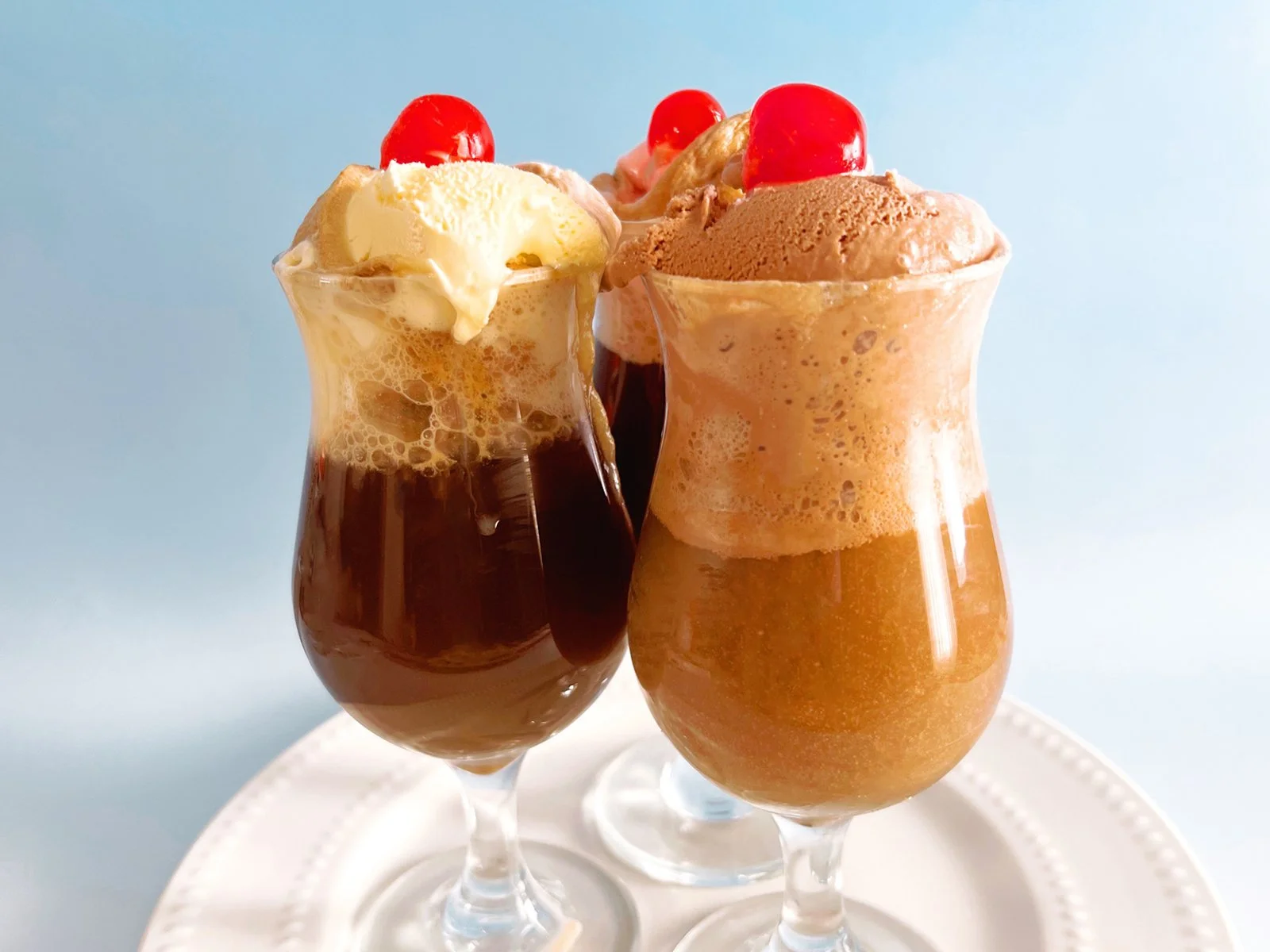 Three retro cola ice cream floats in soda fountain glasses topped with vanilla and chocolate ice cream and maraschino cherries.
