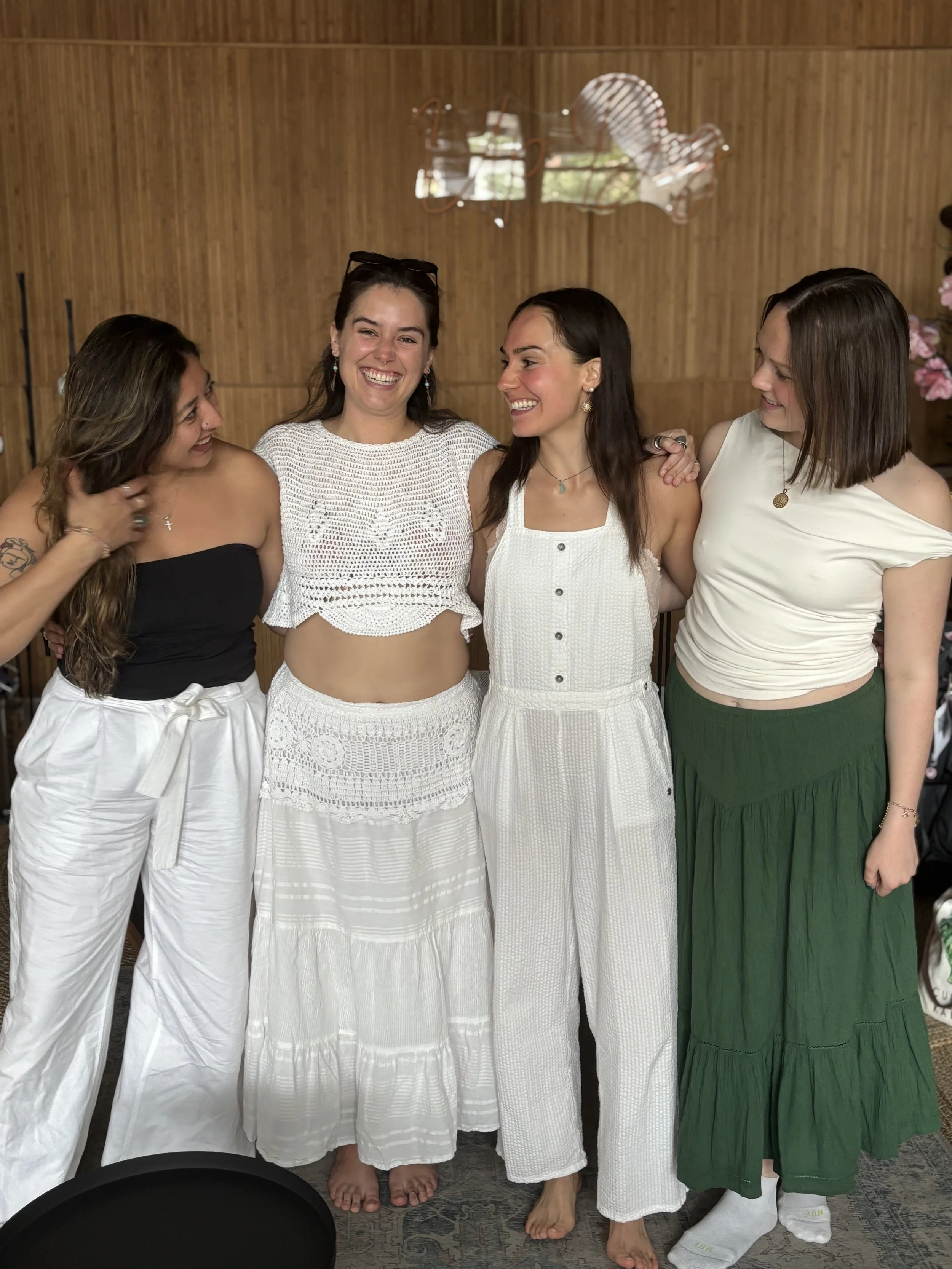 Four women standing together indoors, smiling and looking at each other, with a wooden wall and a mirage-like reflection of chairs in the background.