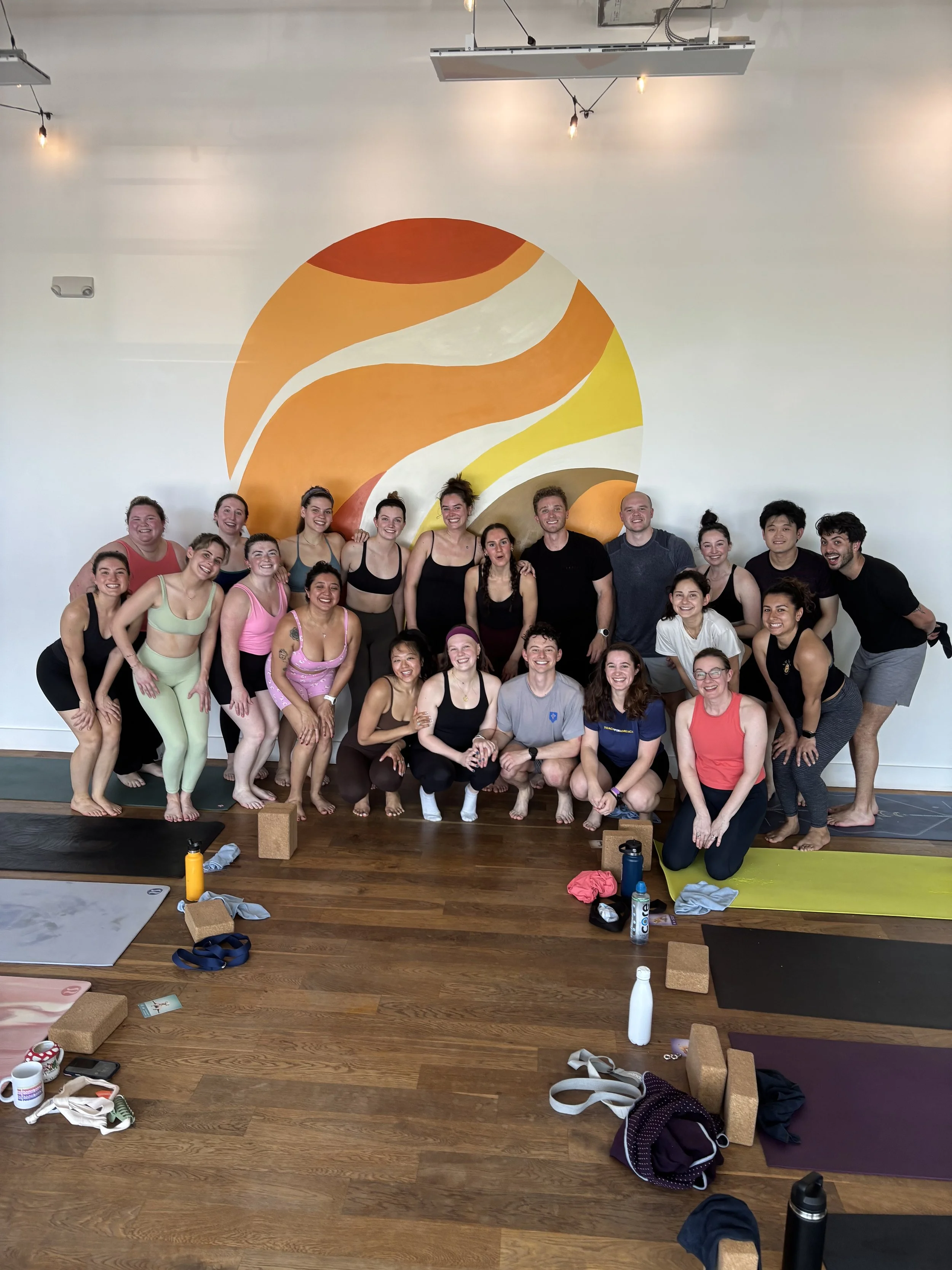 A group of people in workout clothes posing together in a yoga studio with wooden floors and yoga mats, some with water bottles and yoga props around them.