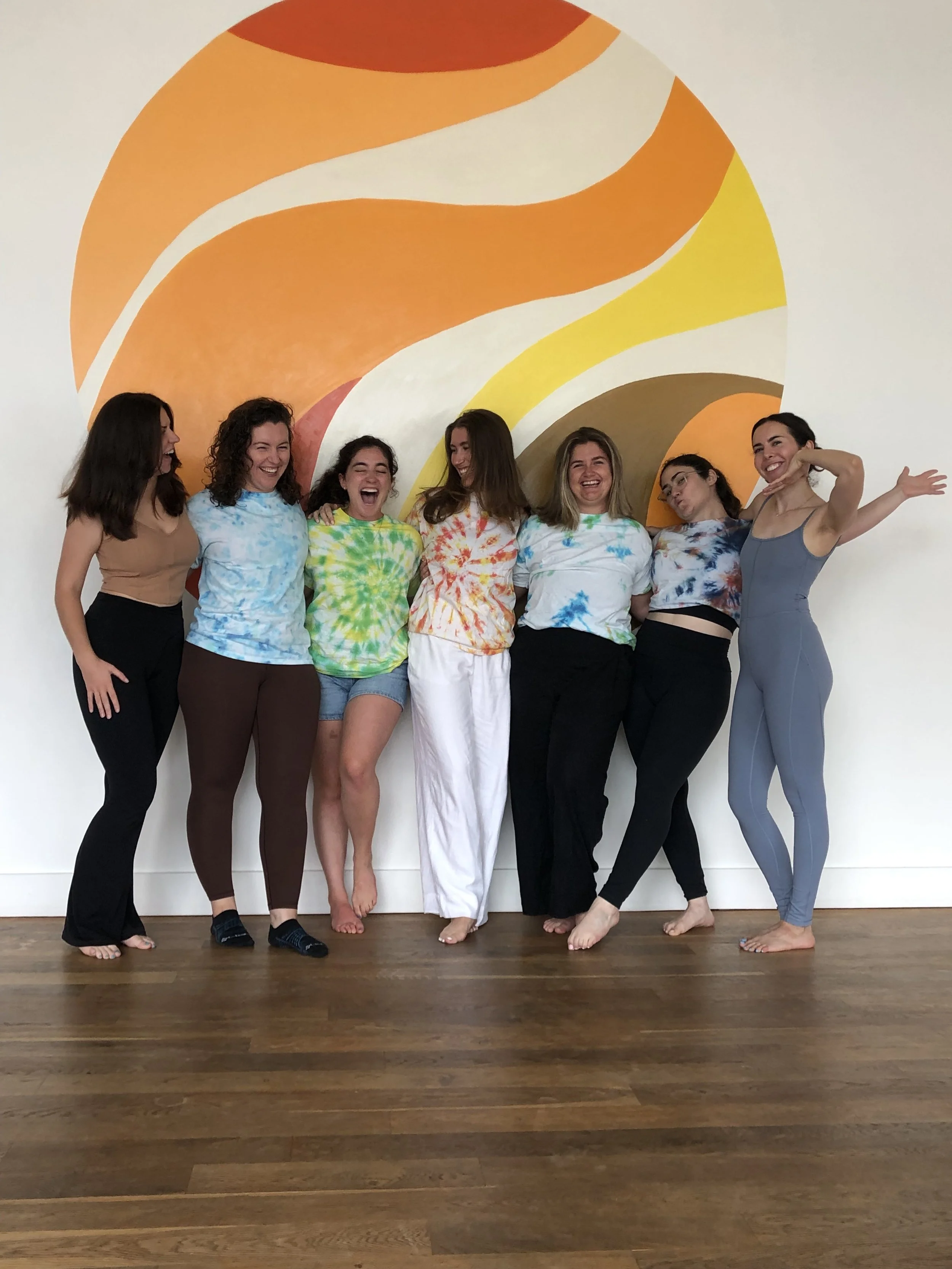 Seven women standing in a row, smiling and laughing indoors in front of a colorful, abstract mural on the wall. They are dressed in casual, tie-dye, and workout clothing.
