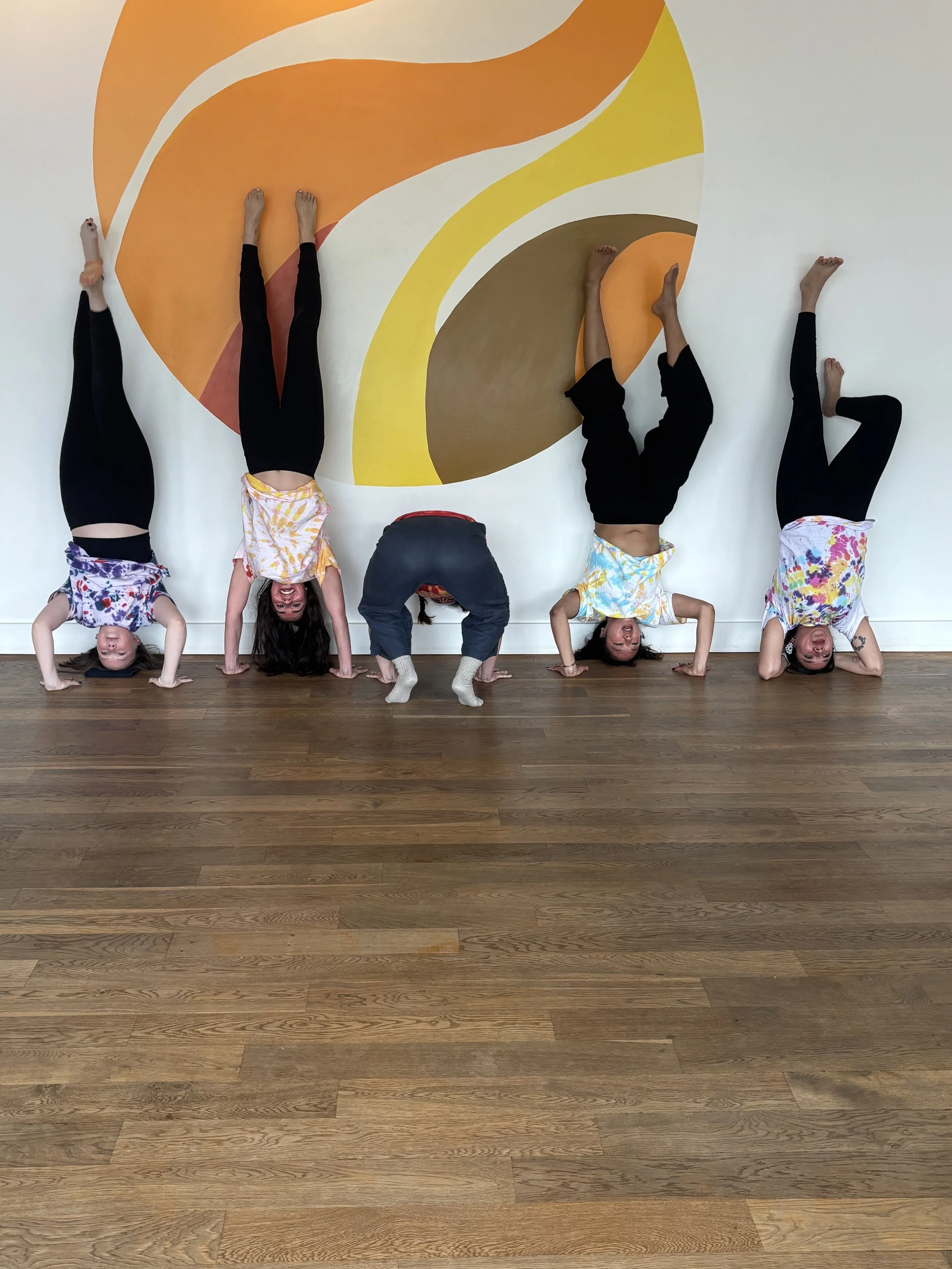Five students in colorful shirts and black pants performing handstands and headstands against a white wall with an abstract orange, yellow, and brown mural.
