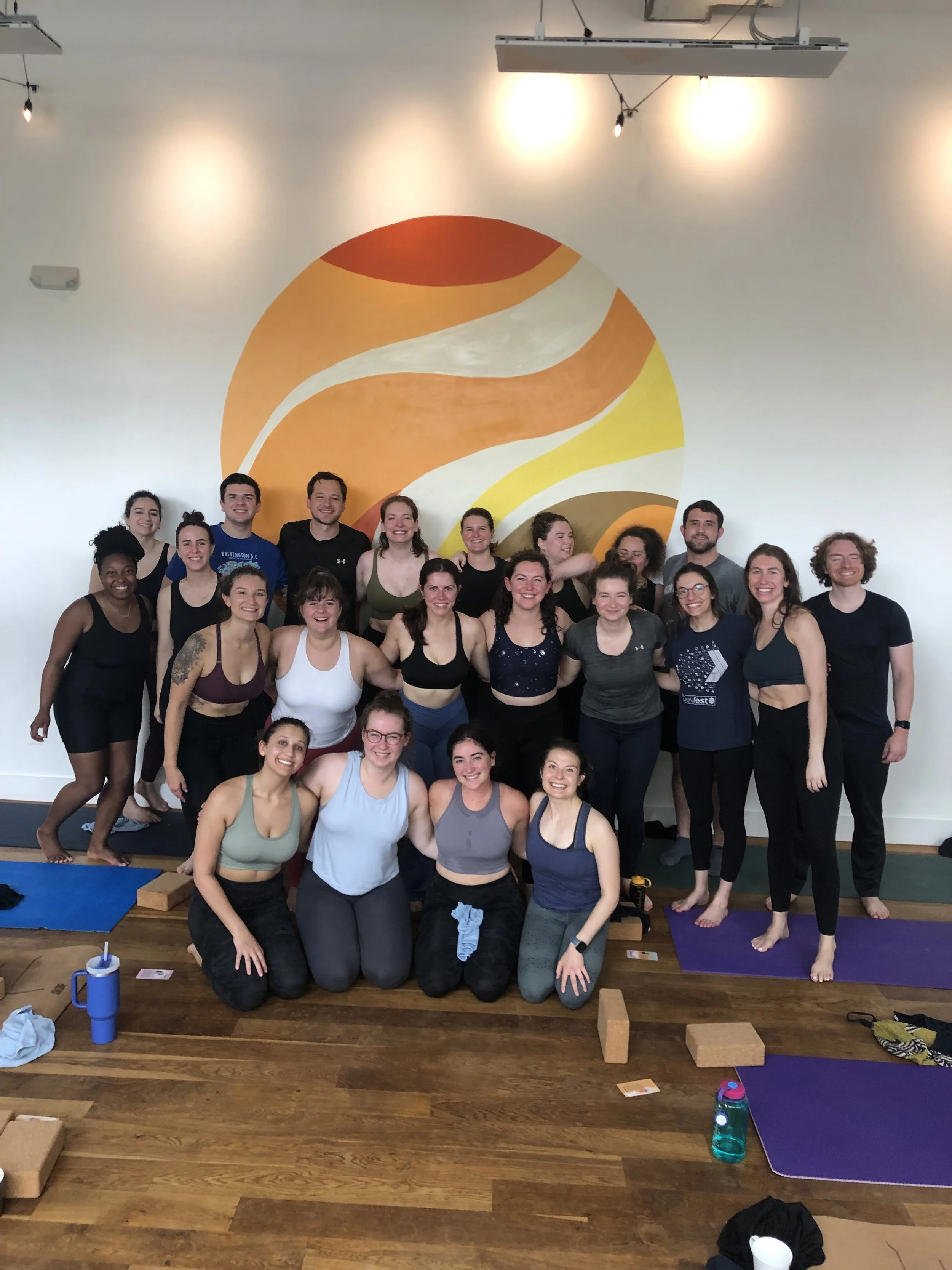 Group of people in a yoga studio with yoga mats, bricks, and water bottles, posing for a photo in front of a wall mural featuring a stylized orange, white, and yellow design.