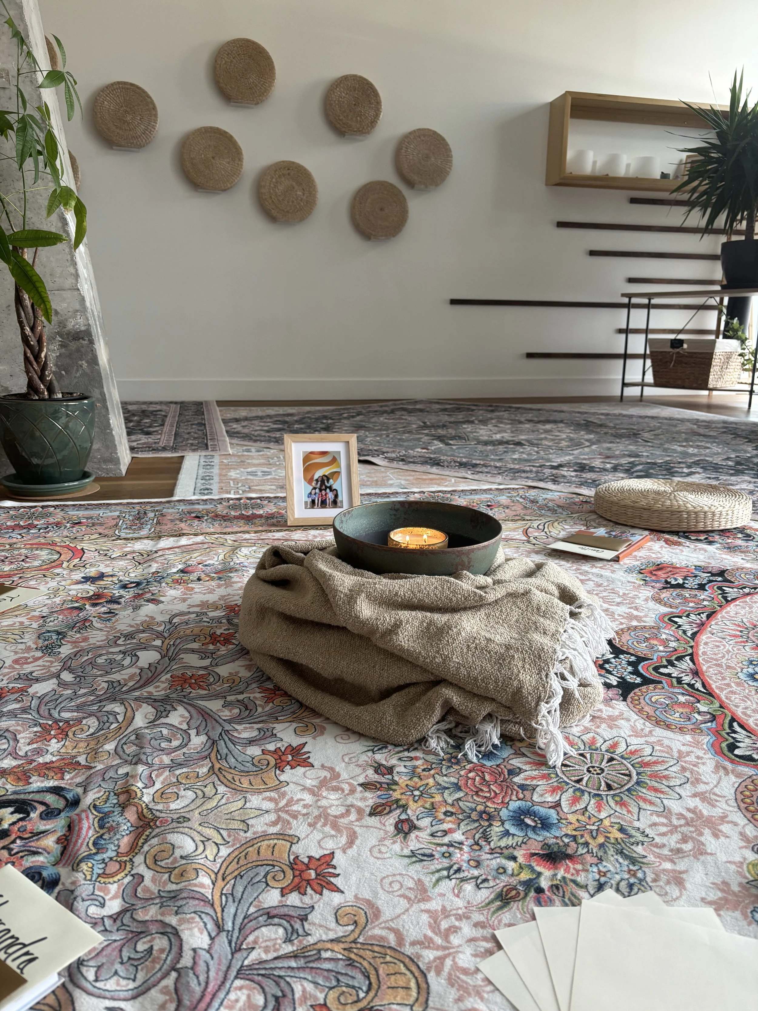 A cozy interior with decorative woven wall hangings, a framed photograph, a lit candle in a bowl, and a layered patterned rug with a blanket and a book on it.