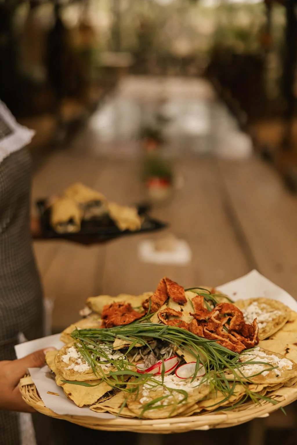 A hand holding a basket of traditional Mexican tacos topped with herbs, vegetables, and shredded meat, with a blurred background of a marketplace or restaurant setting.