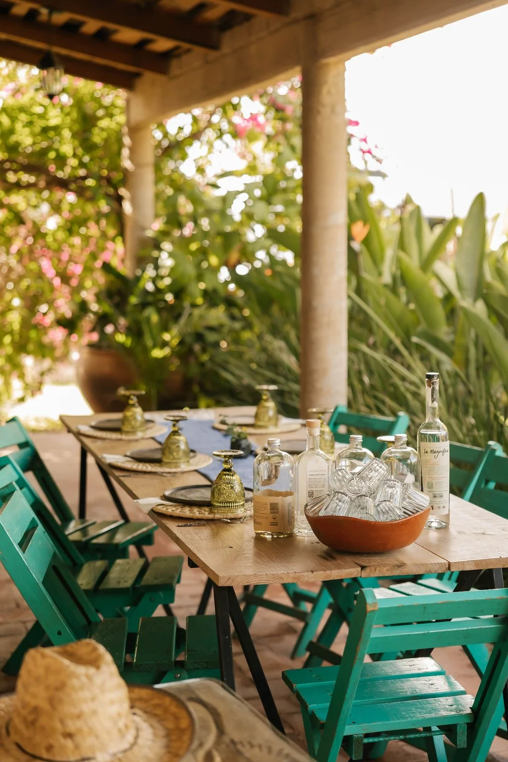 An outdoor dining table set for a meal, with turquoise chairs, glass bottles, glasses, and a large bowl on a wooden table surrounded by lush green plants during a private mezcal experience. 