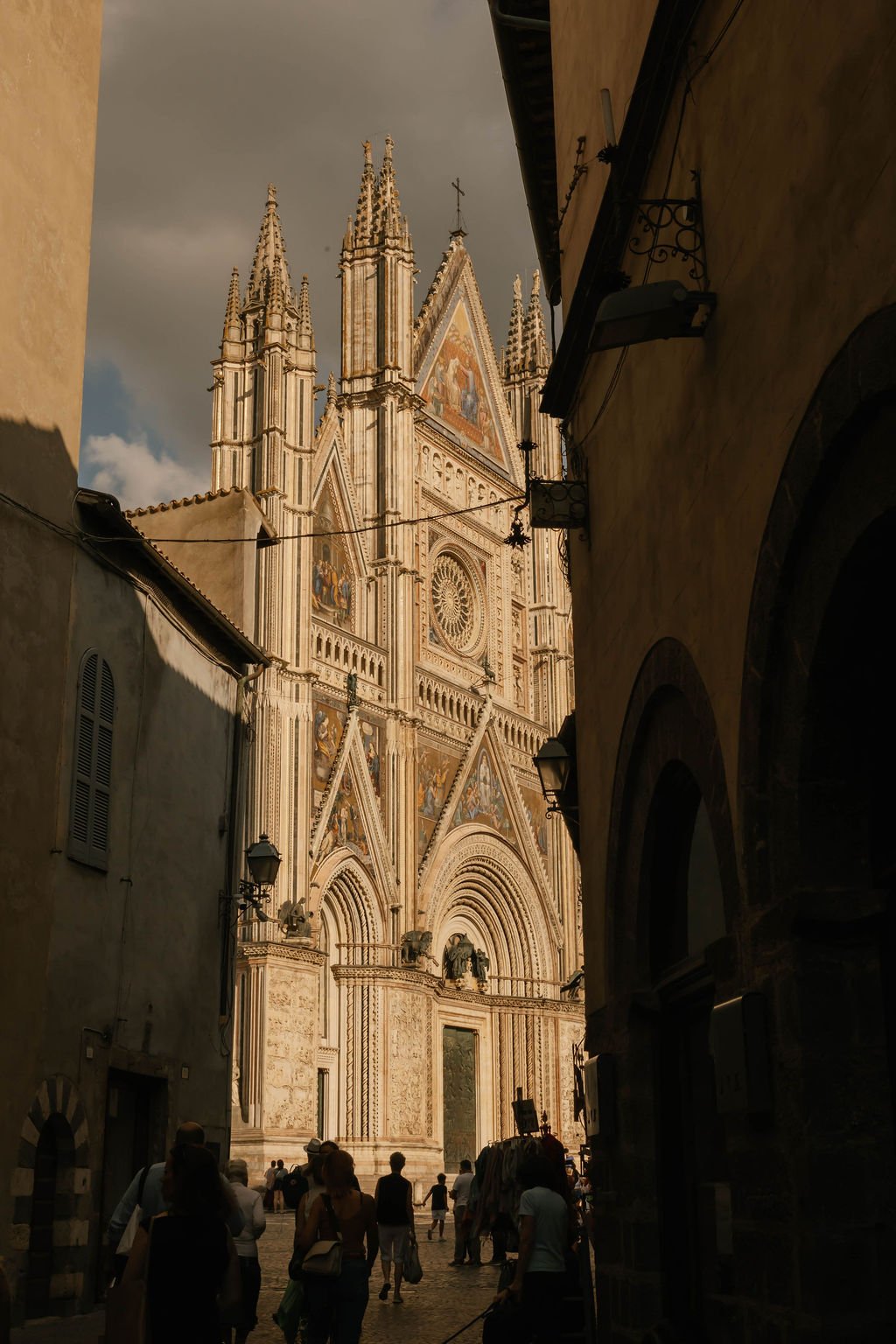View of the Siena Cathedral in Italy with intricate Gothic architecture, spires, and detailed façade, seen from a narrow street with people walking.