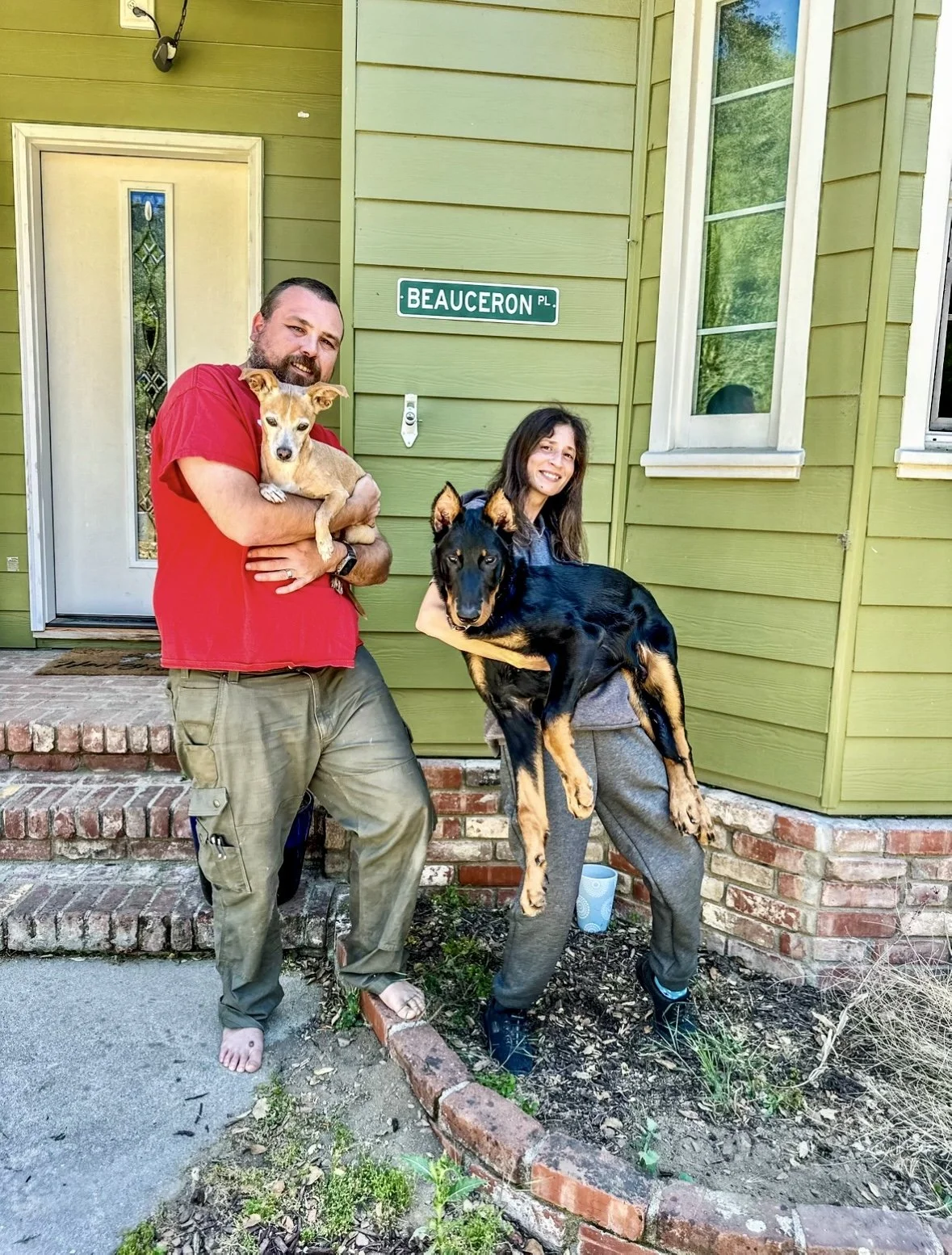A man, woman, and two dogs standing outside in front of a green house with a street sign that reads 'Beauceron Pl.' The man has a beard, is barefoot, wearing a red shirt and cargo pants, and is holding a small tan dog. The woman has long dark hair, is smiling, wearing a gray hoodie and sweatpants, and is holding a large black and tan dog. The scene takes place on a brick step and a small garden bed with dirt and plants is visible.
