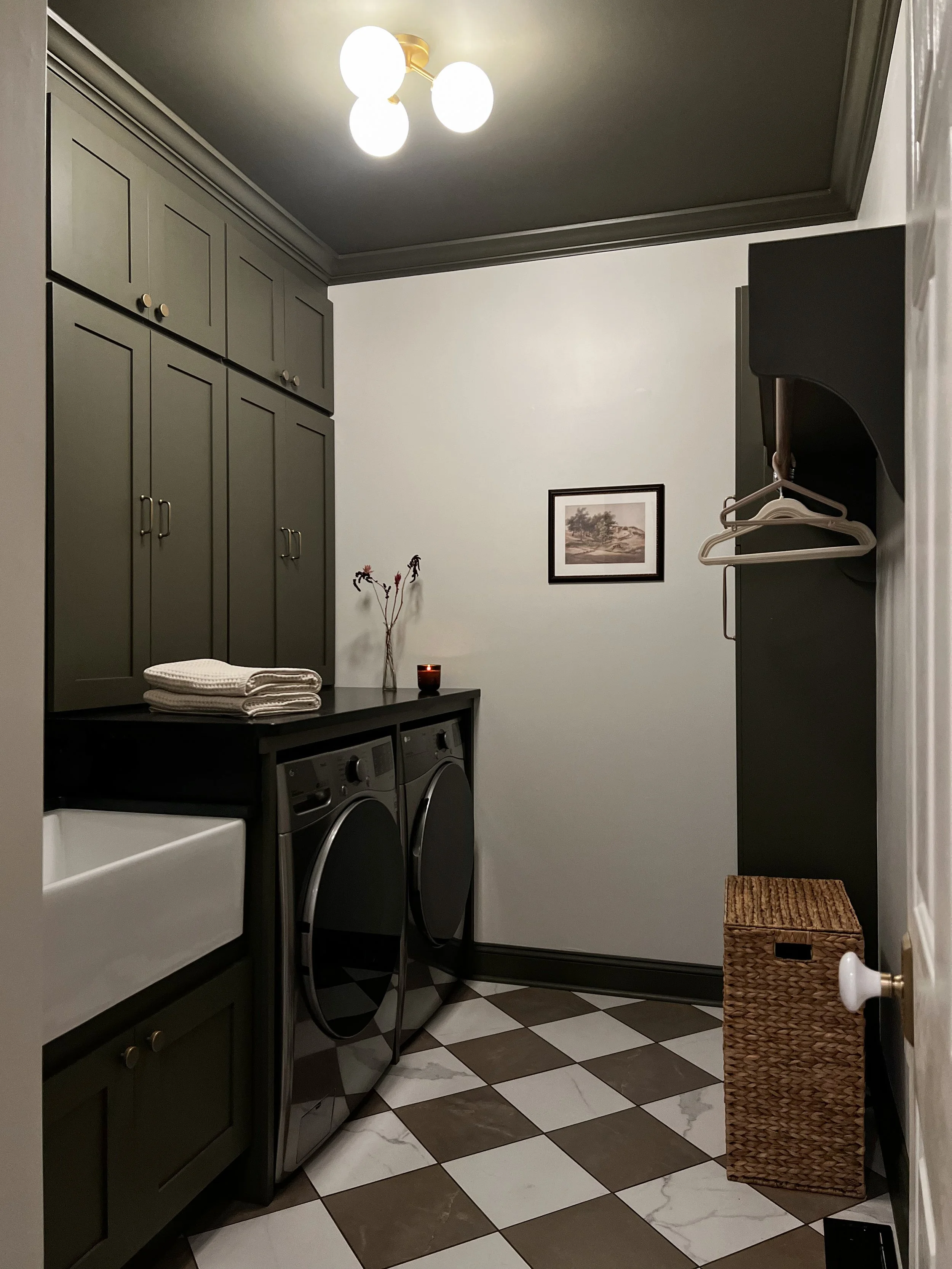 Interior design of an upscale laundry room for a Philadelphia area home. Embodying a modern yet timeless style, this space features dark olive custom cabinets, black granite counter above built-in laundry machines, and checker tile flooring.