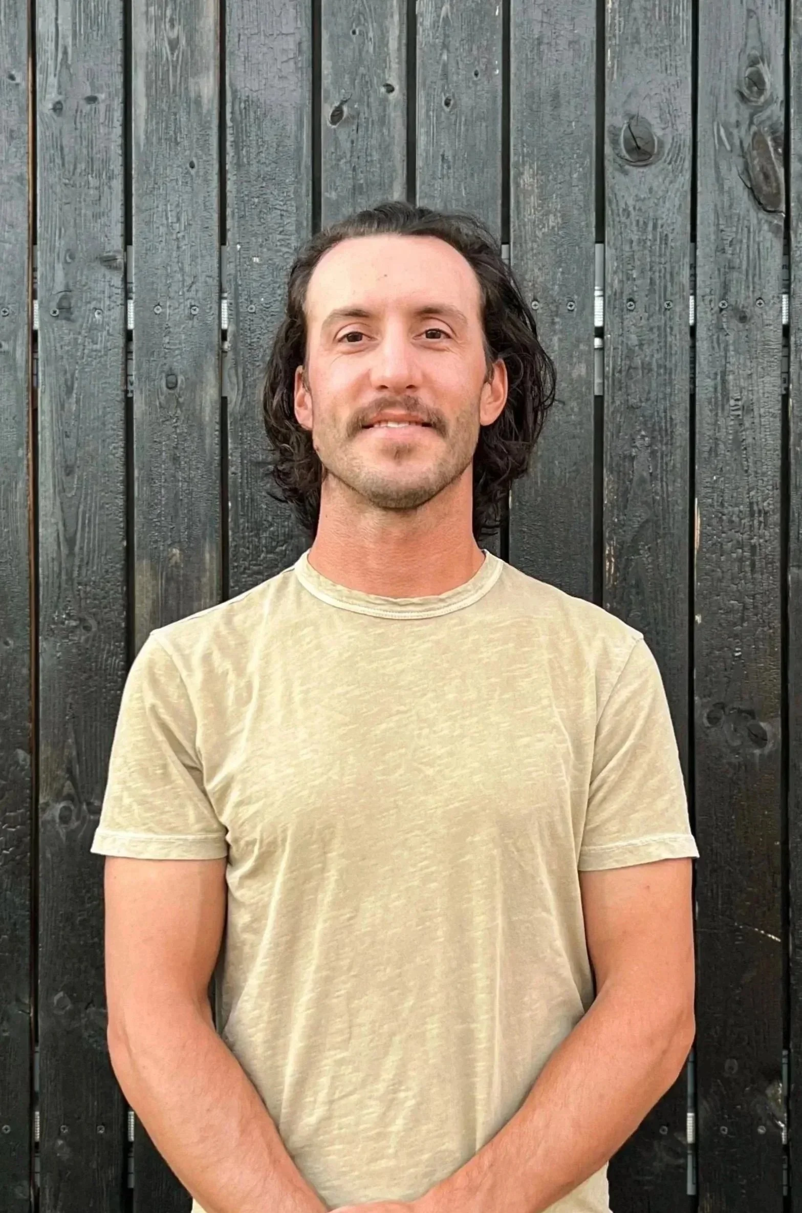 A man with medium-length wavy dark hair, a mustache and goatee, wearing a beige t-shirt, standing in front of a black wooden fence.