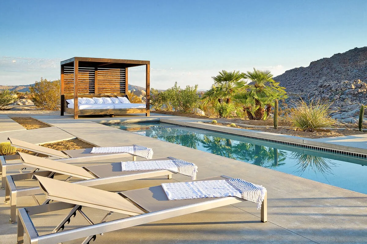 Desert backyard with swimming pool, lounge chairs with towels, a cabana with cushions, desert plants, cacti, and mountains in the background under a clear blue sky.