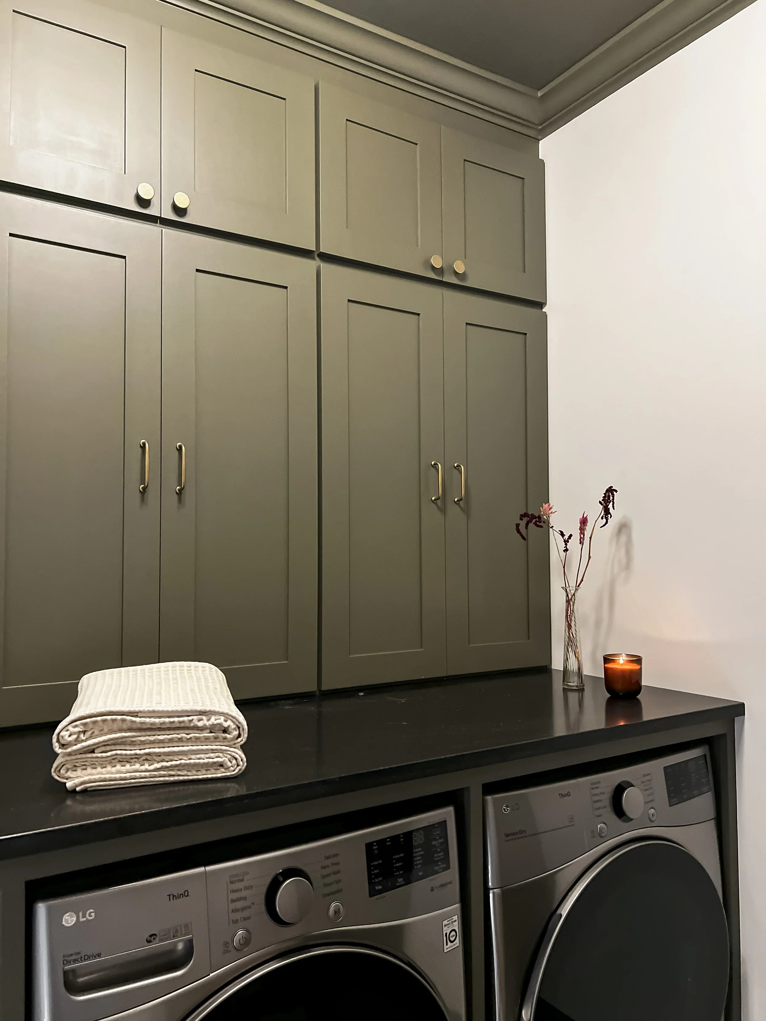 Interior design of an upscale laundry room for a Philadelphia area home. Embodying a modern yet timeless style, this space features dark olive custom cabinets, black granite counter above built-in laundry machines, and checker tile flooring.