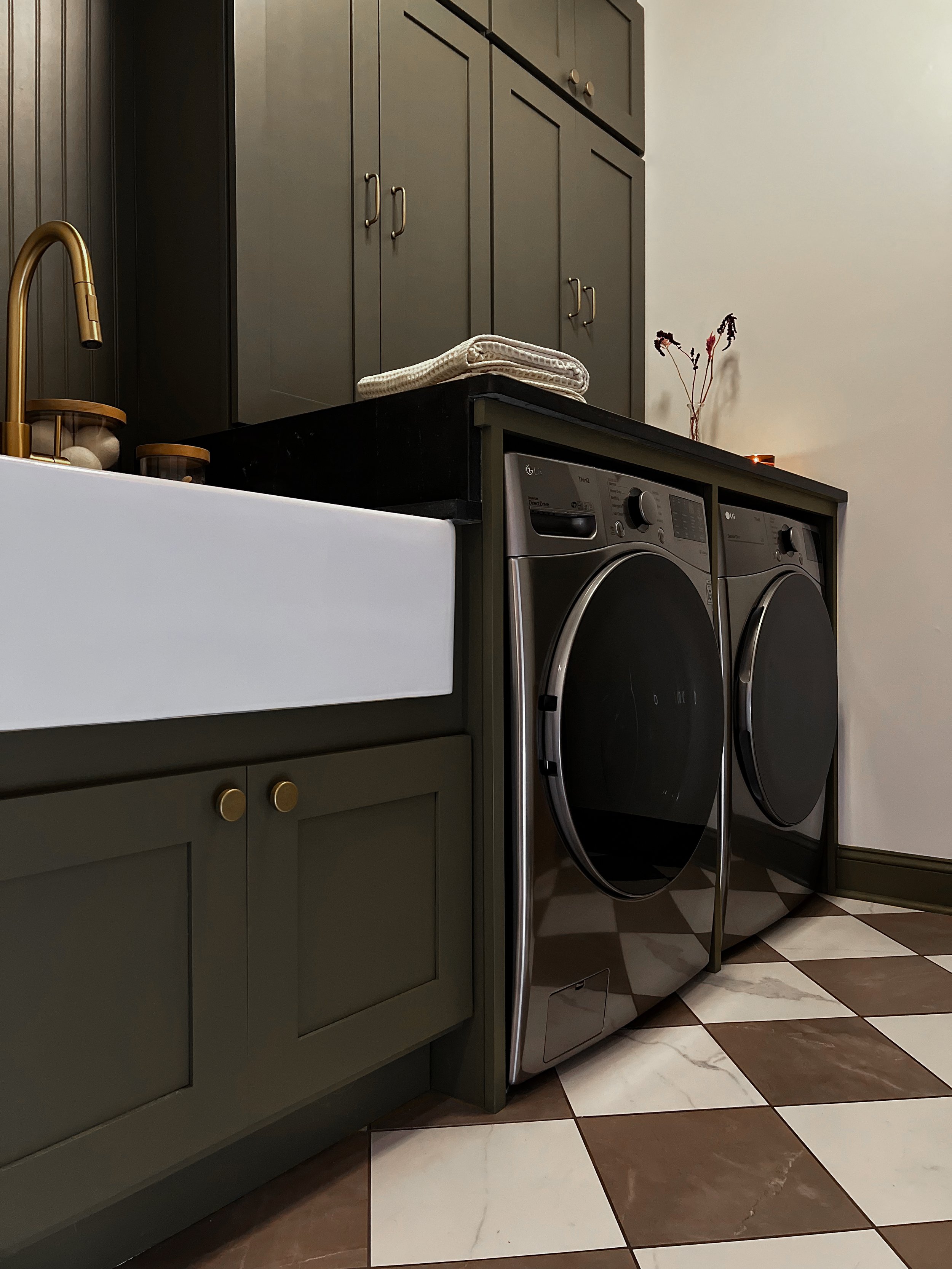 Interior design of an upscale laundry room for a Philadelphia area home. Embodying a modern yet timeless style, this space features dark olive custom cabinets, black granite counter above built-in laundry machines, and checker tile flooring.