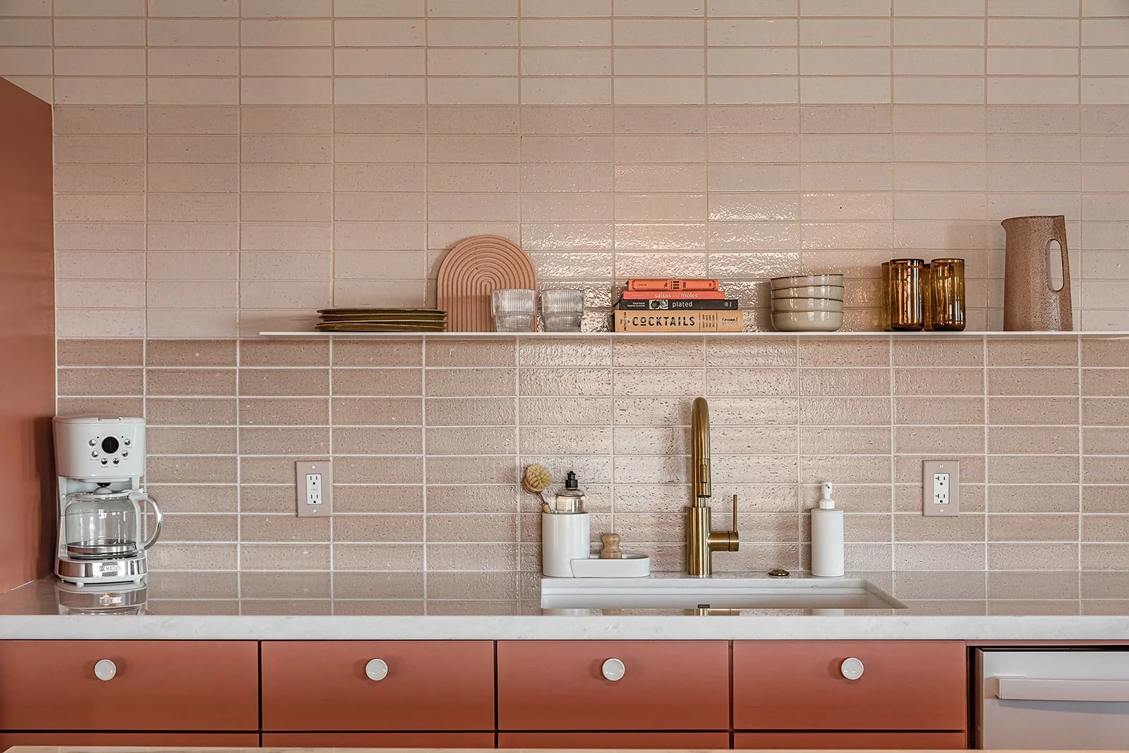 Kitchen design with custom pink cabinets, blush backsplash tile wall, white quartzite counter, sleek metal floating shelf with brass fixtures.