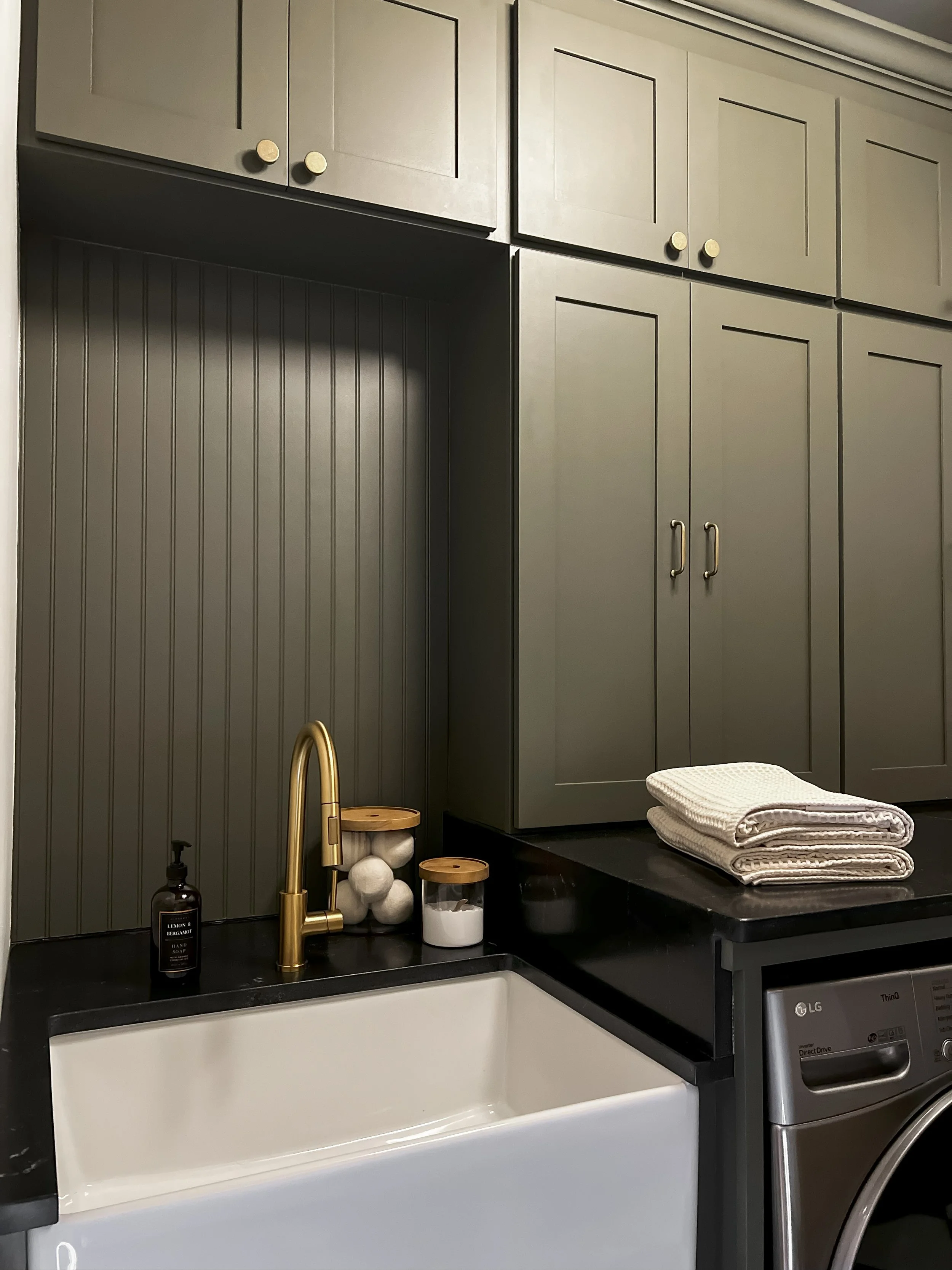 Interior design of an upscale laundry room for a Philadelphia area home. Embodying a modern yet timeless style, this space features dark olive custom cabinets, black granite counter above built-in laundry machines, and checker tile flooring.