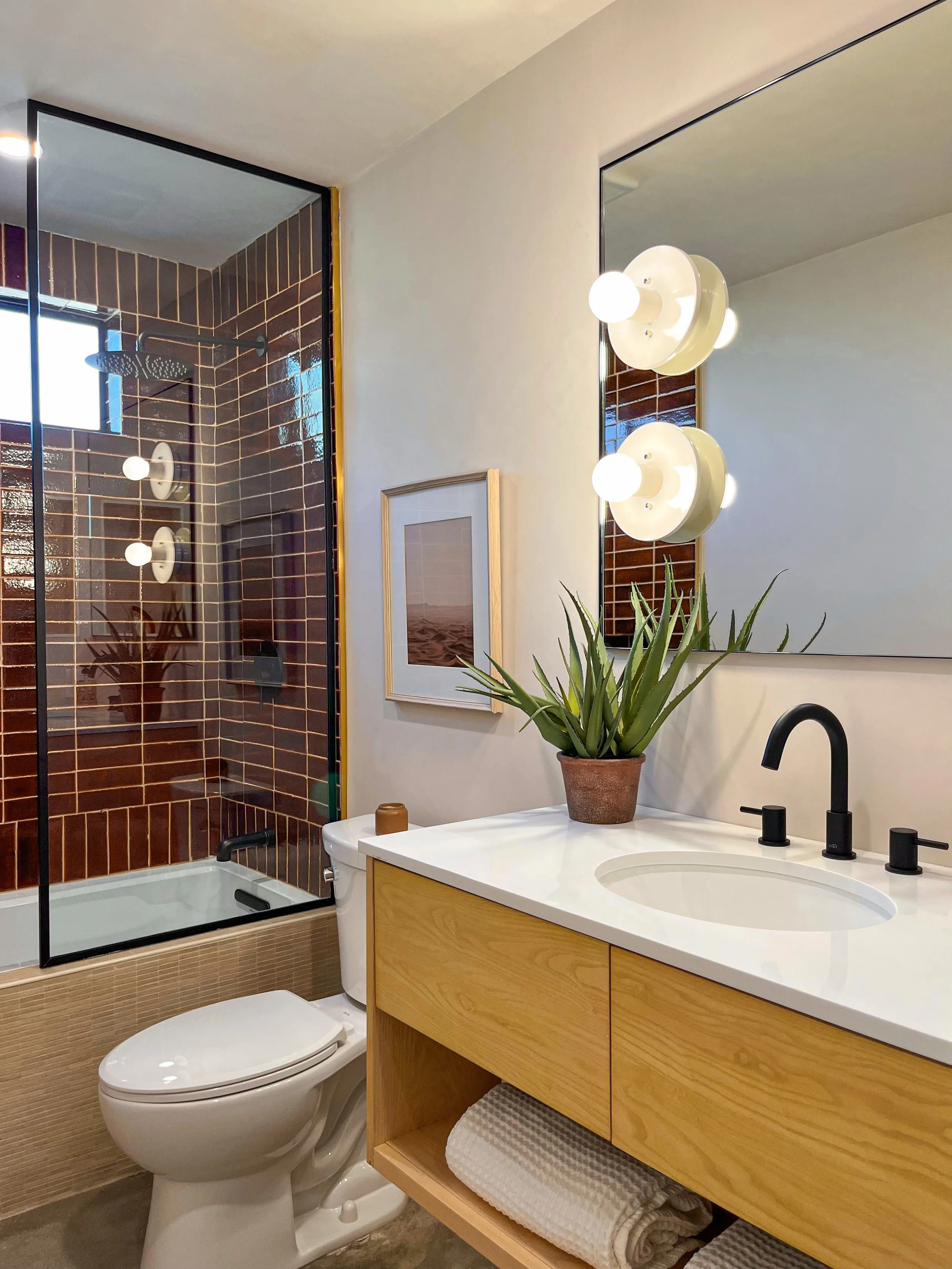 Modern bathroom with brown tile shower, a wood vanity with a white countertop, a potted plant, a large mirror, and a framed picture on the wall.