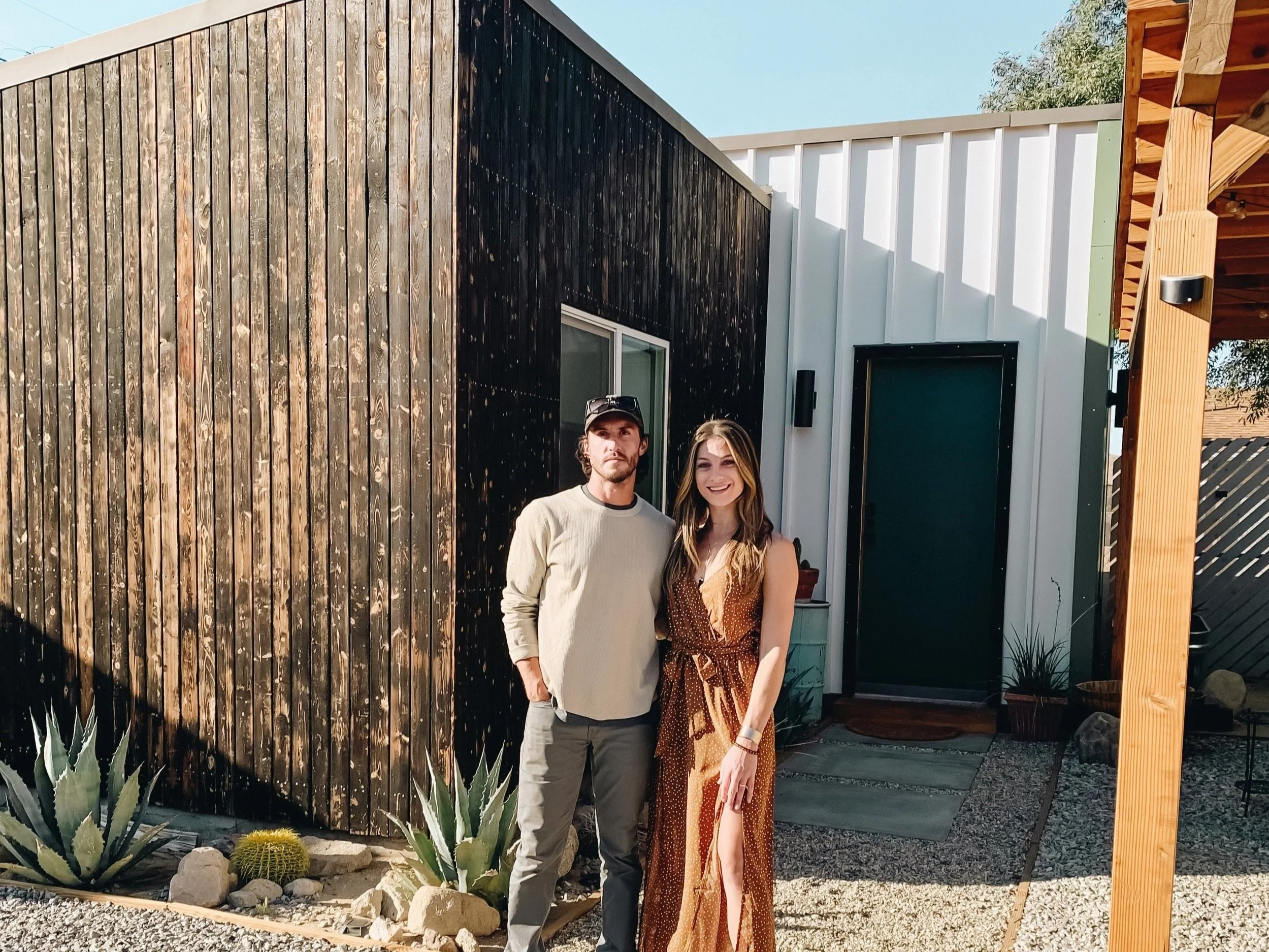 A man and woman standing outside a modern house with a black wooden and white metal exterior, cactus plants, and a gravel yard during daytime.