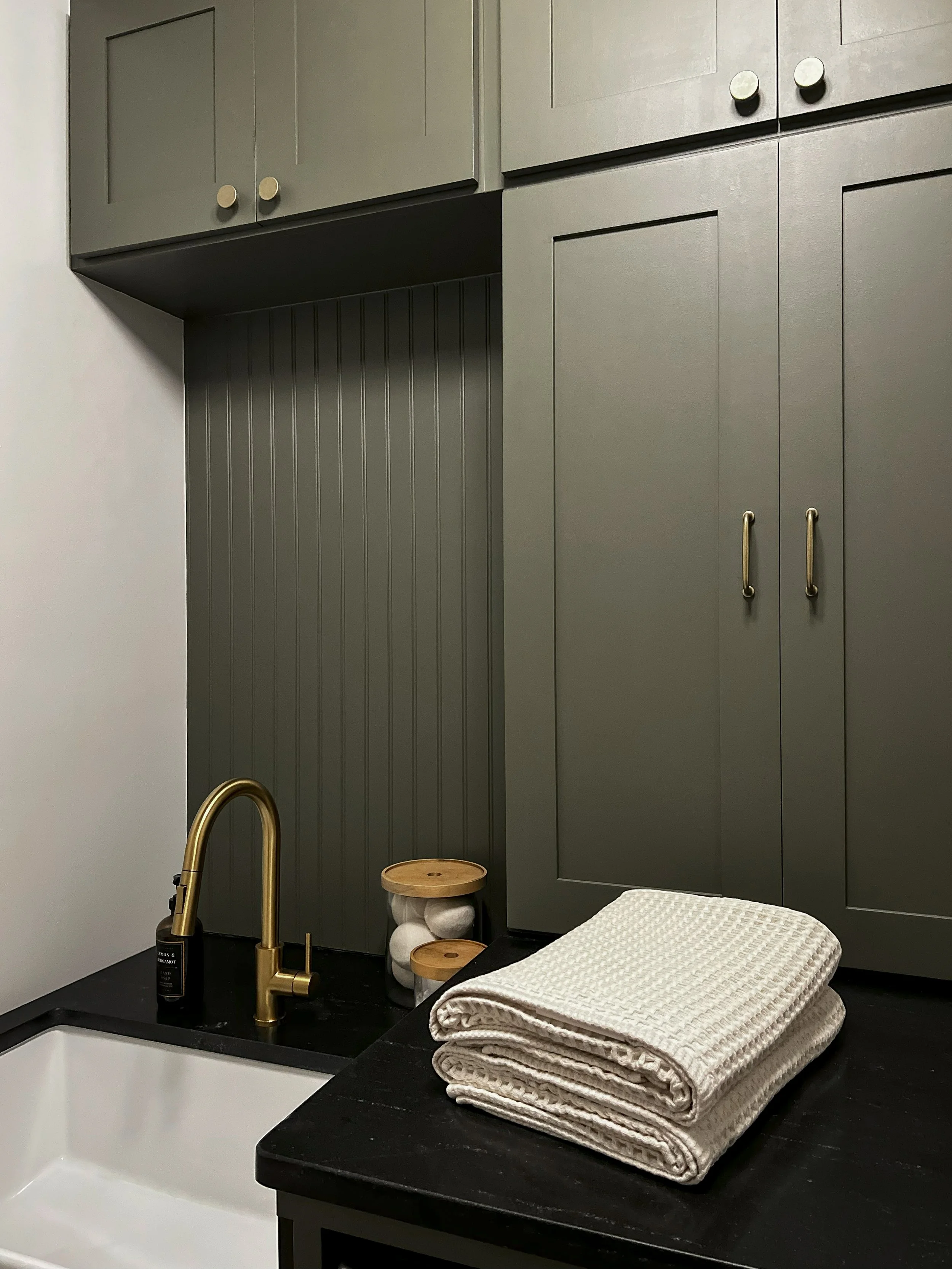 Interior design of an upscale laundry room for a Philadelphia area home. Embodying a modern yet timeless style, this space features dark olive custom cabinets, black granite counter above built-in laundry machines, and checker tile flooring.